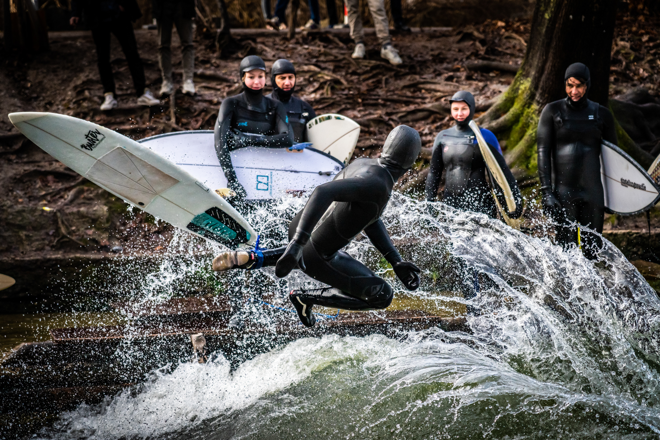 Group of people in black wetsuits with white surfboards beside rushing water and rocks in wooded area.