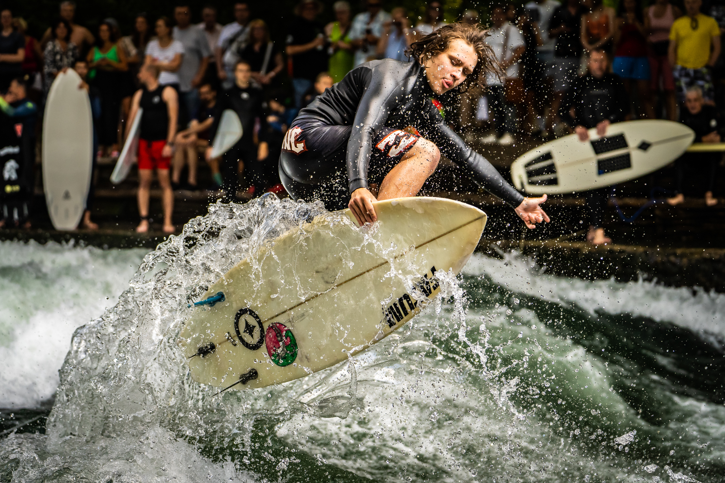 Surfer in black wetsuit on white surfboard creating splash in green water, with crowd watching in background.