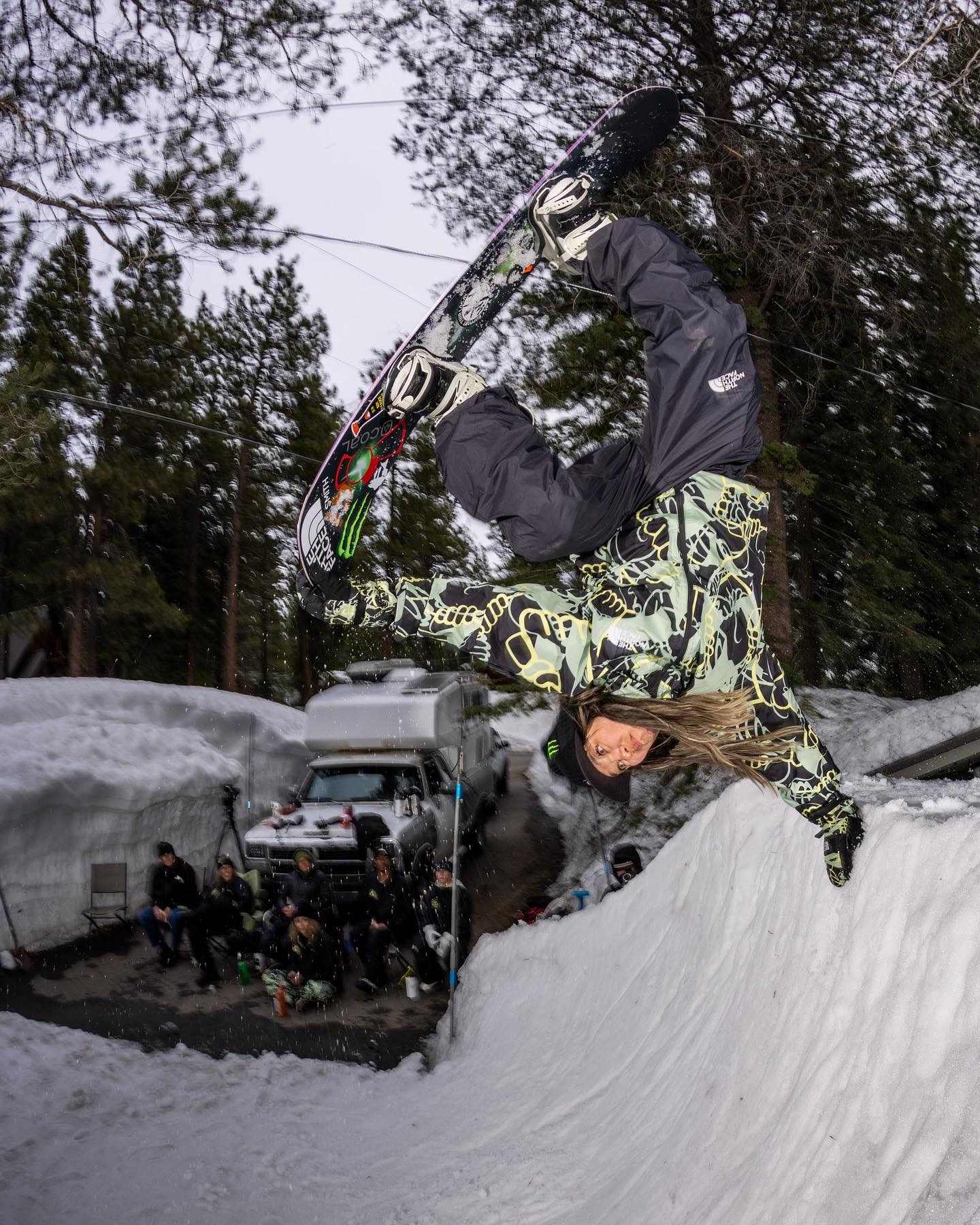 Snowboarder in grey jacket and patterned trousers performs aerial trick above snowy halfpipe, surrounded by evergreen trees.