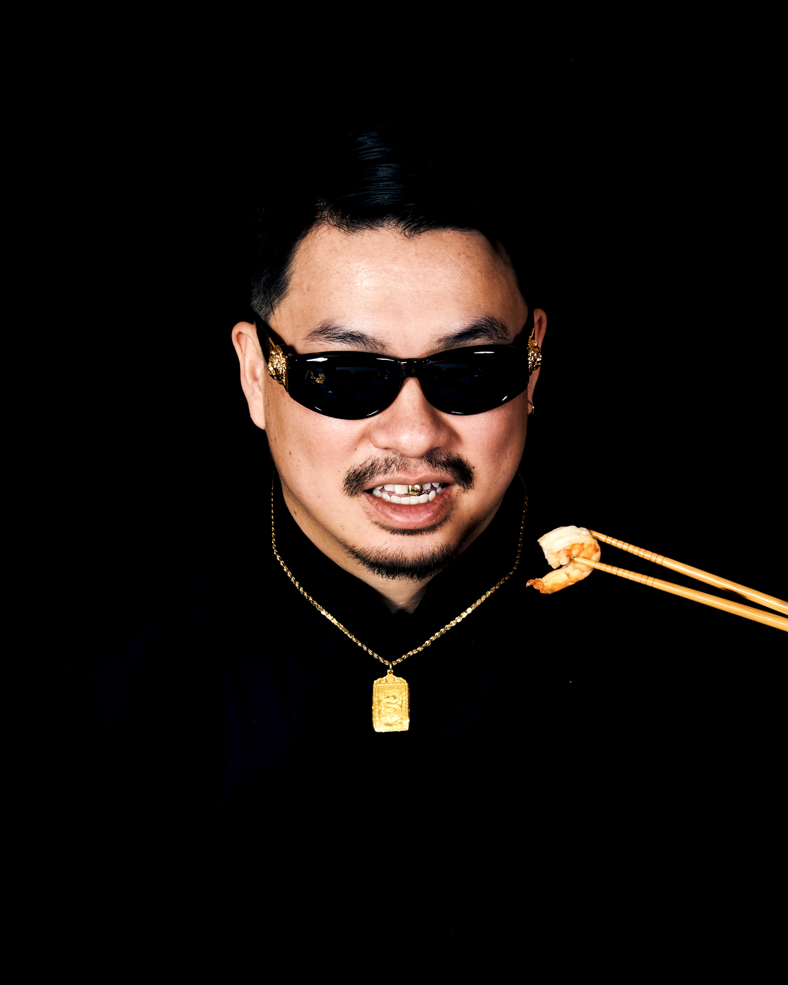 Man wearing black sunglasses and gold chain necklace with pendant, holding chopsticks with food, against black background.