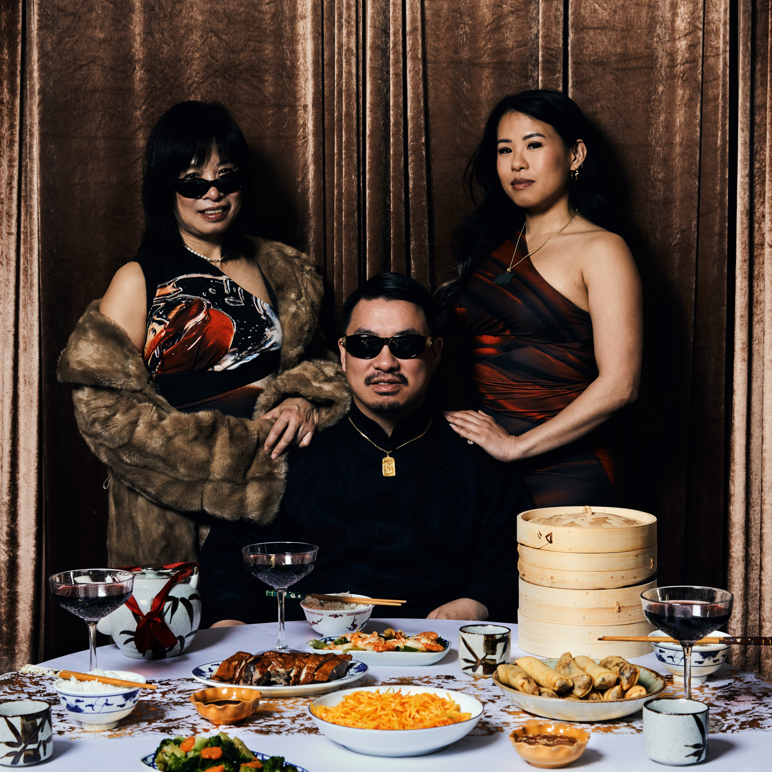 Three people wearing sunglasses posed behind table with Chinese dishes, bamboo steamer, and wine glasses against ornate wooden backdrop.