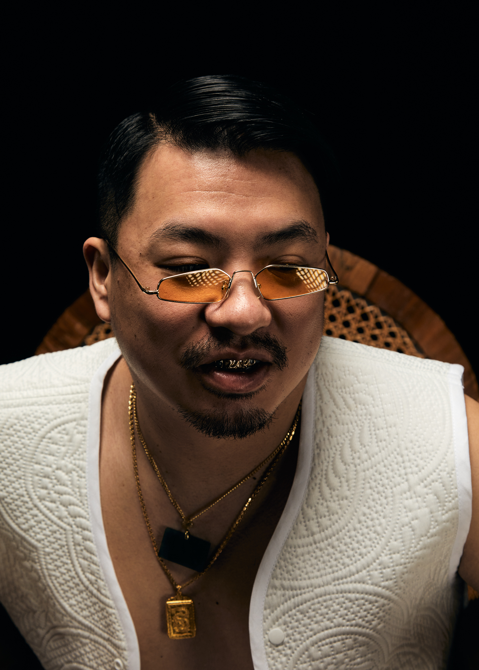 Man with orange-tinted glasses, gold chains, and white textured jacket against black background. Dark hair slicked back, goatee.