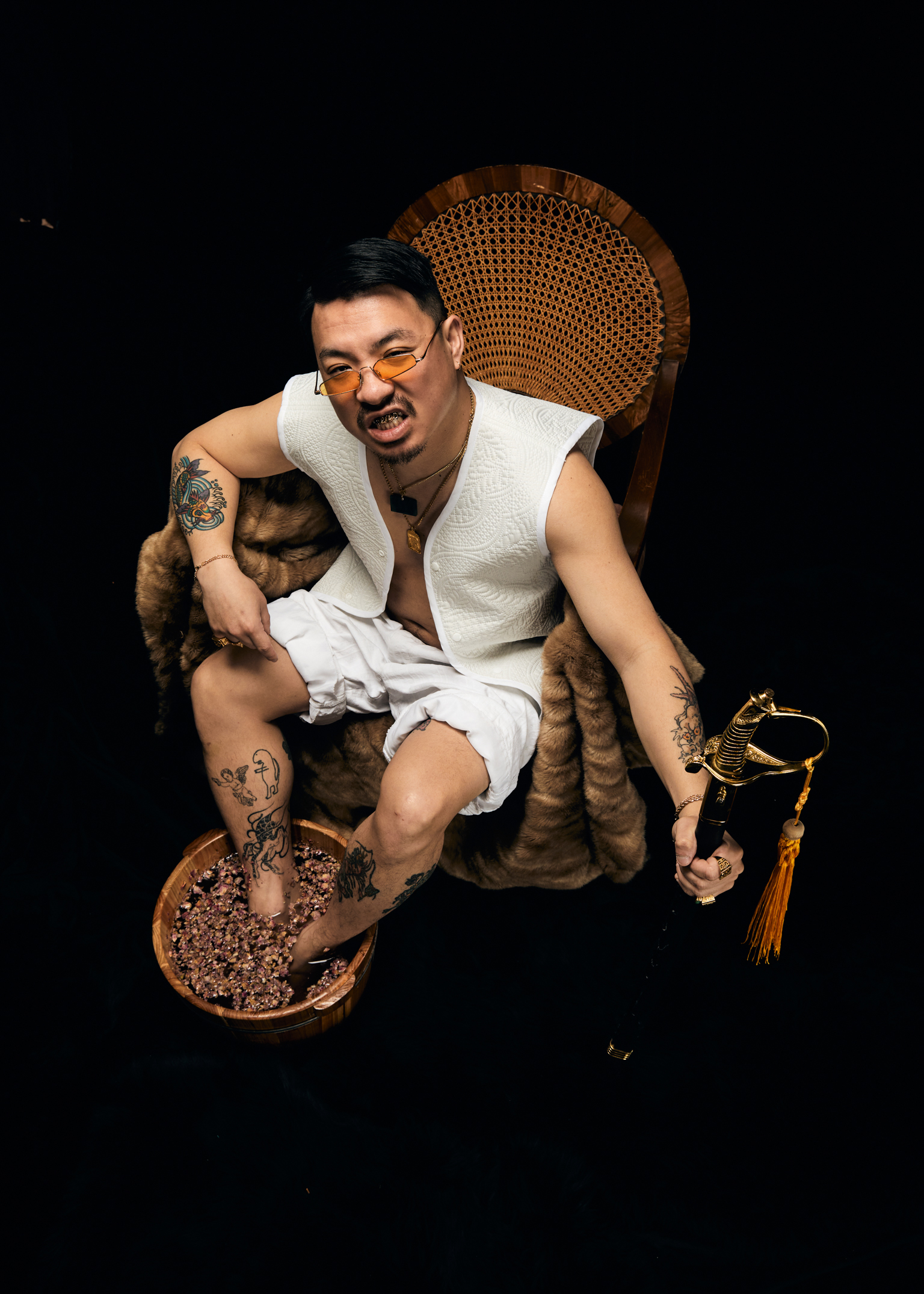 Man with tattoos sits in wicker peacock chair holding tabby cat, wearing white vest and shorts against black background.