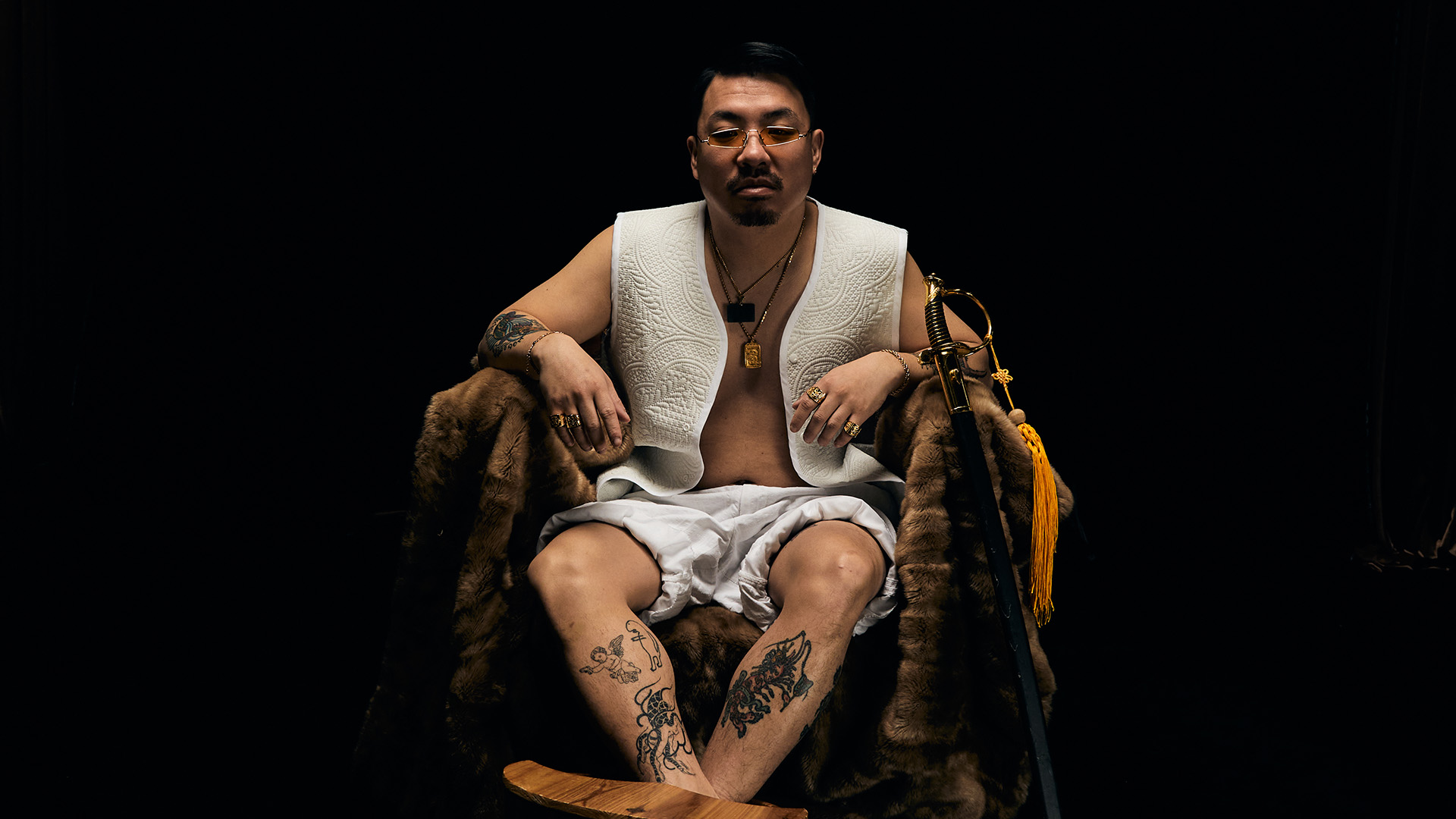 Asian man with moustache sitting in wooden chair against black background, wearing white waistcoat and shorts, tattoos on legs.