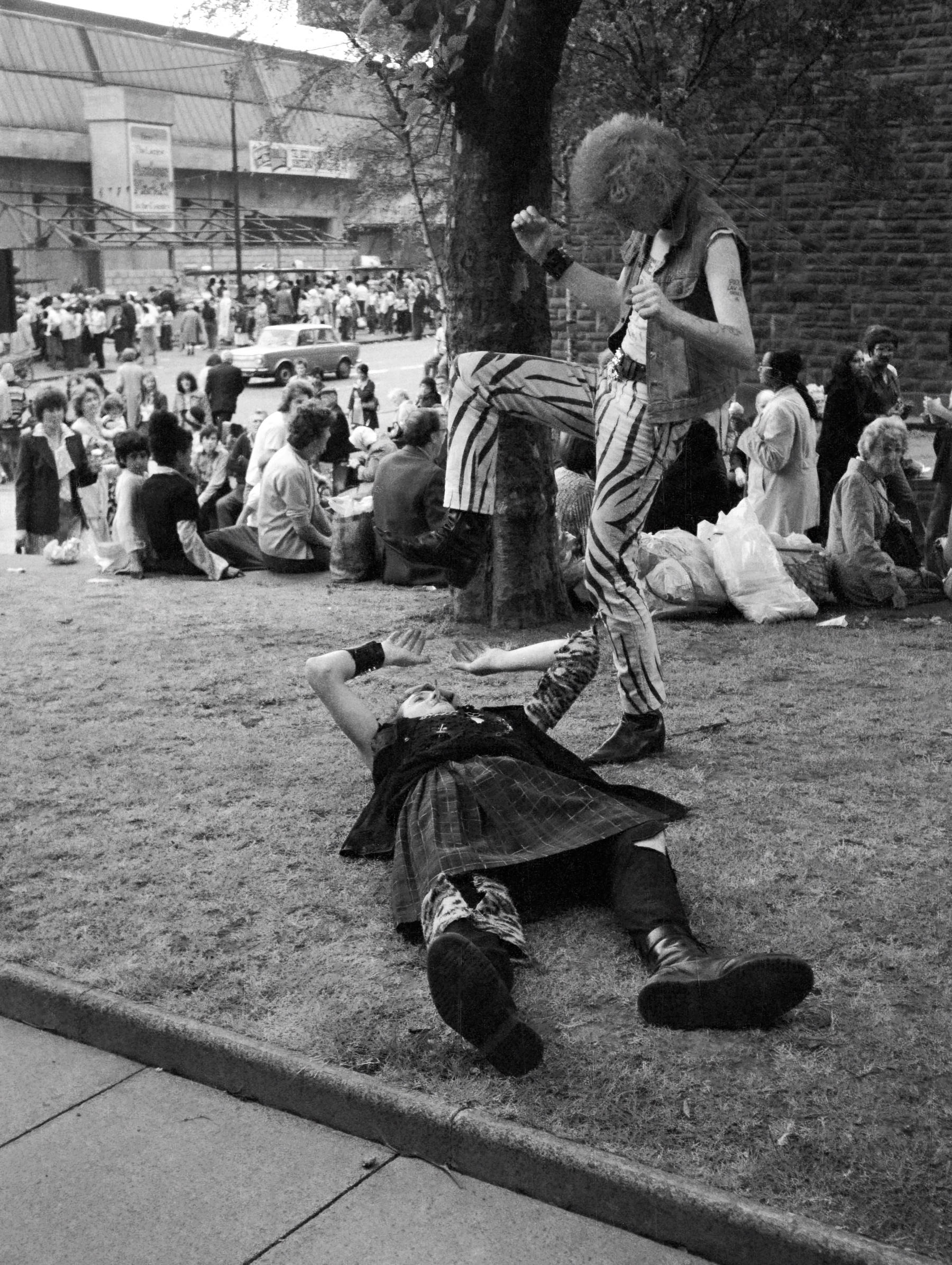 Black and white street scene showing performer in striped trousers standing over person lying on pavement, with crowd gathered around tree and buildings in background.