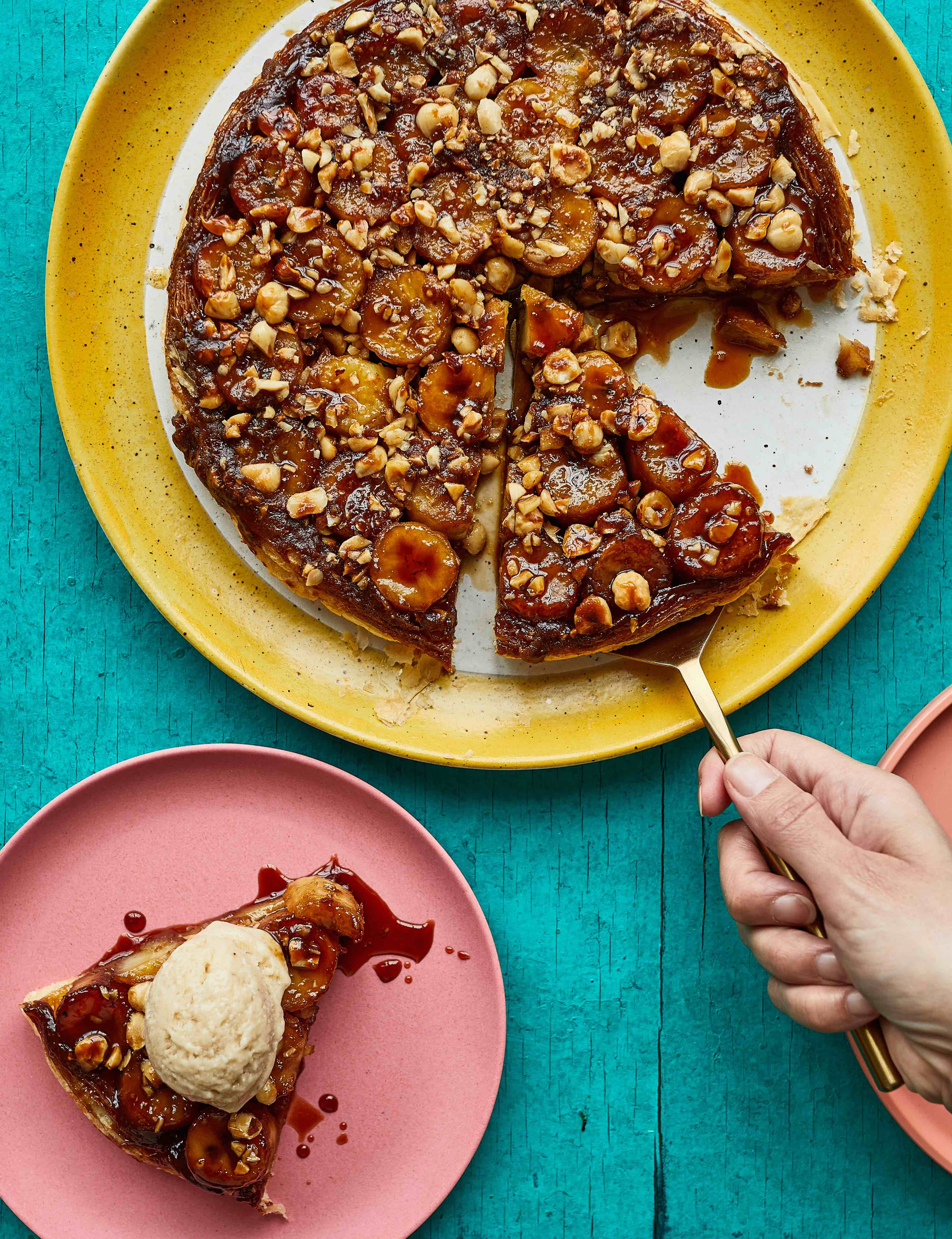 Dark chocolate tart topped with nuts and dried fruit on yellow plate, with slice on pink plate topped with cream. Hand holds fork.