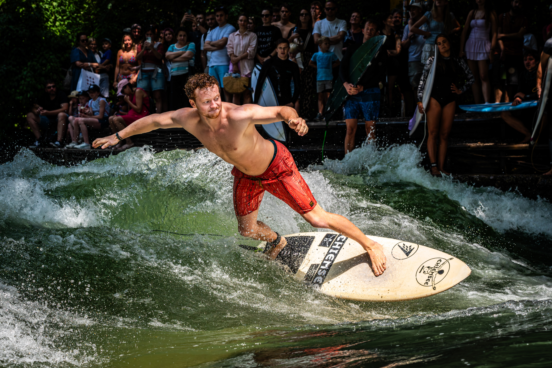 Shirtless man in red shorts surfing on white board in green water with crowd watching from background.