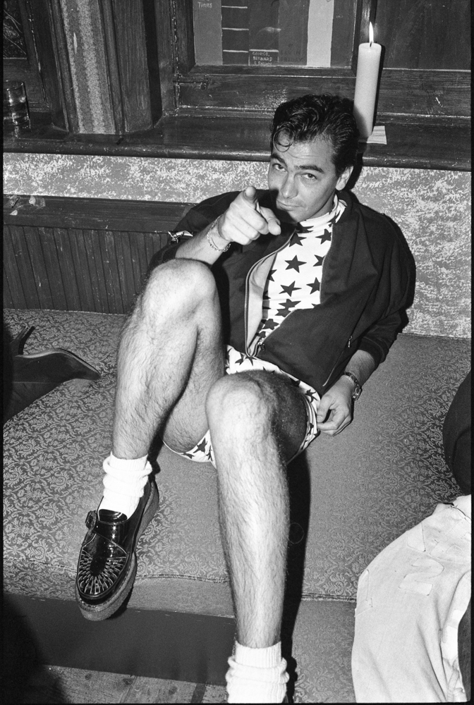 Black and white image of man sitting on steps wearing star-patterned vest, dark jacket, shorts, white socks and lace-up shoes.