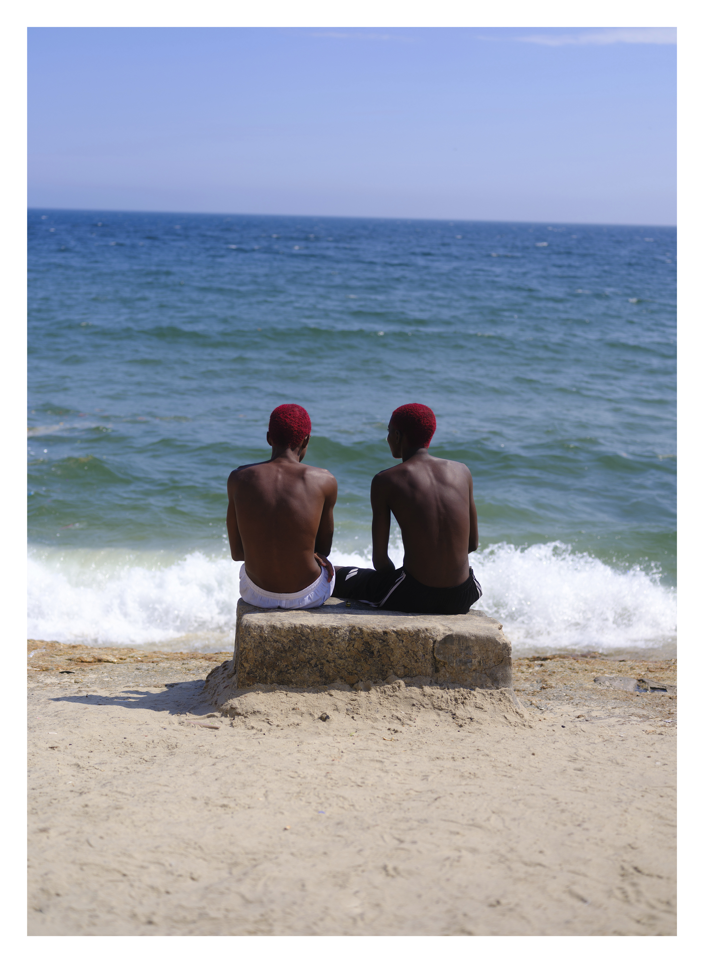 Two shirtless men in red caps sitting on concrete block at water's edge, turquoise sea with white foam, sandy beach, blue sky.