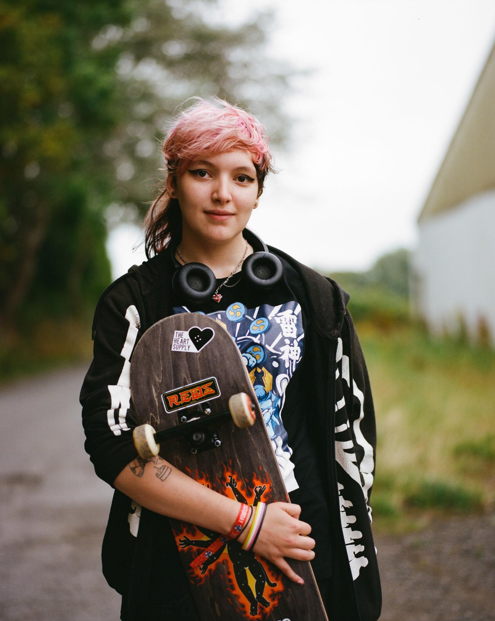 Young woman with pink hair wearing a patterned jacket, standing in a wooded area.