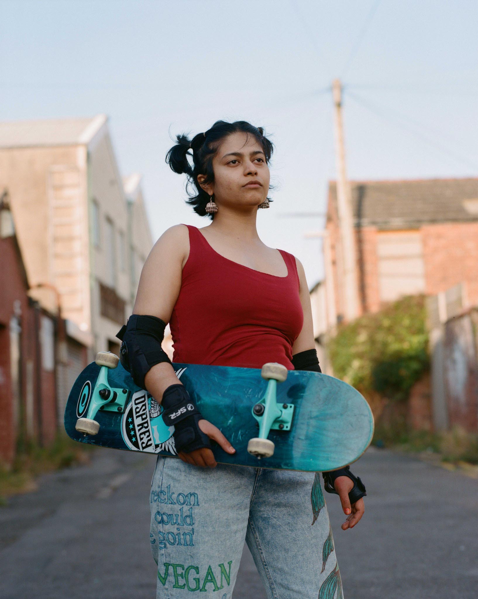 Woman in red top holding a skateboard, standing on a paved road with buildings in the background.