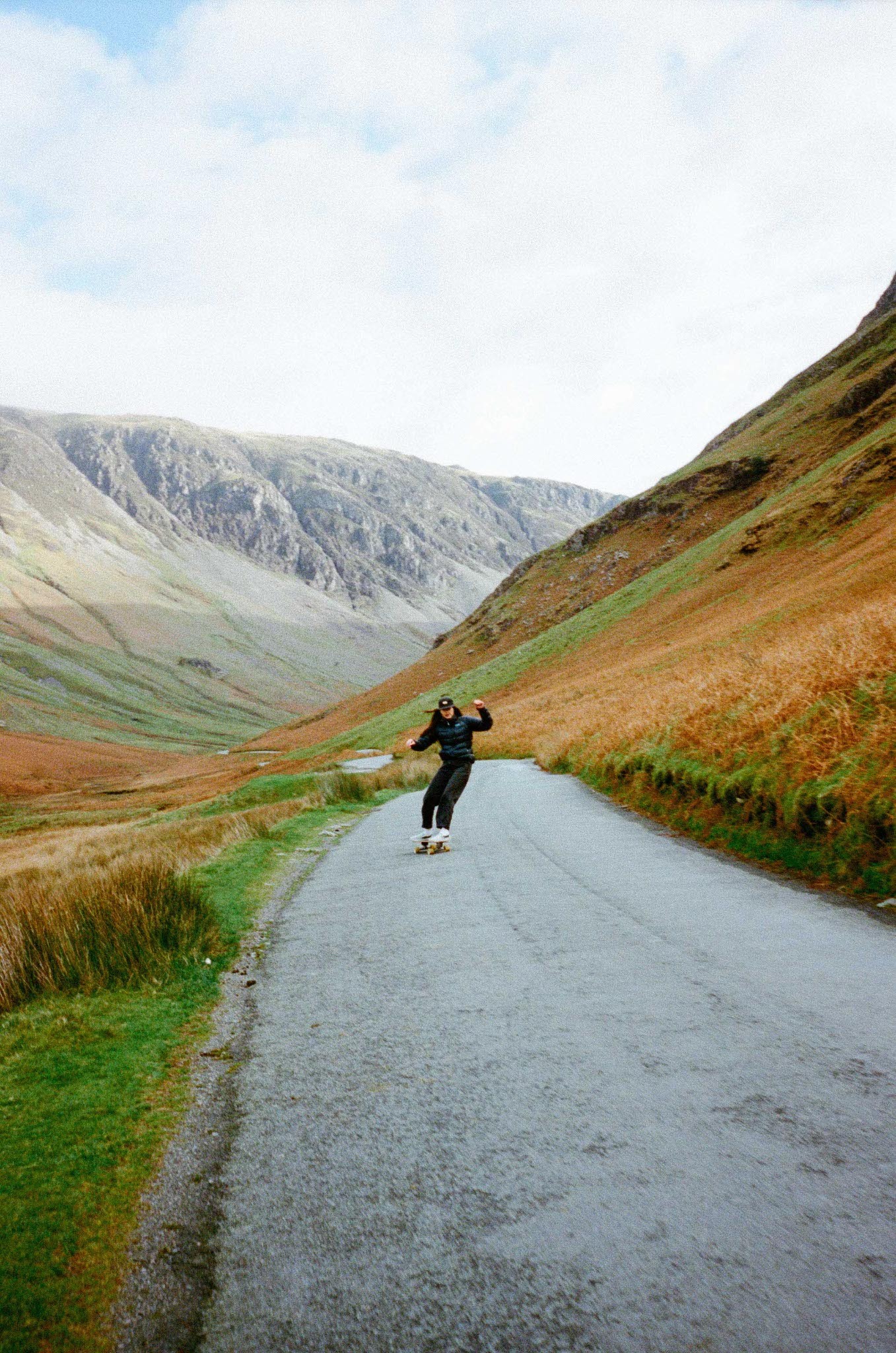 Winding road through mountainous, grassy landscape with person on skateboard.