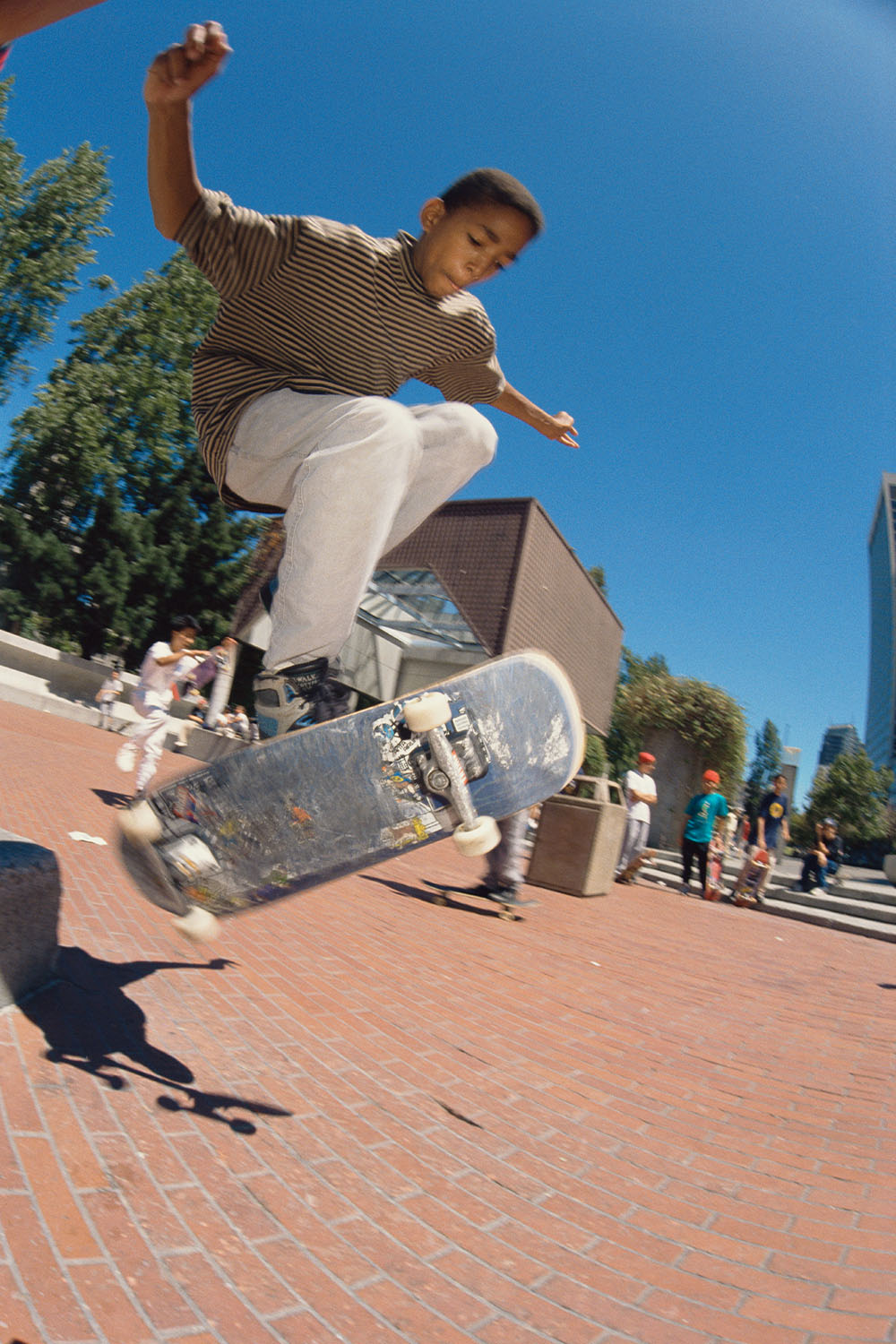 Skateboarder in striped shirt and white trousers performing trick on skateboard above brick plaza, blue sky and trees behind.