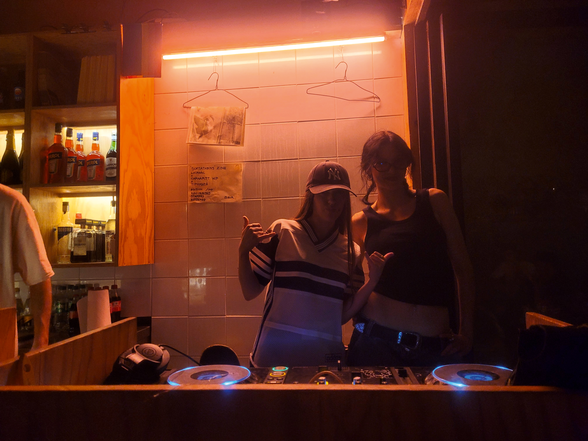 Two people in kitchen with warm orange lighting from overhead strips. Dark foreground with blue-lit surfaces, shelving visible left side.