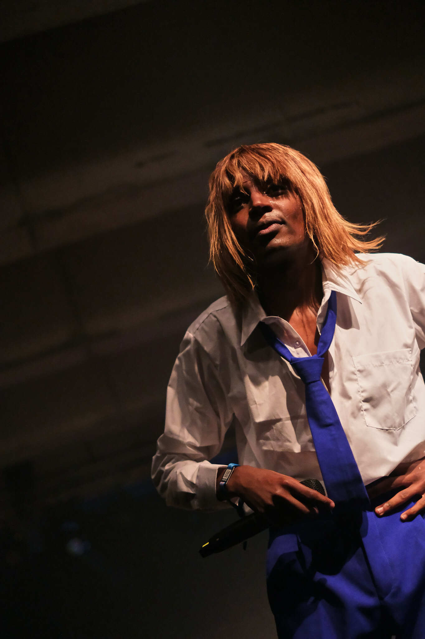 Person wearing a white shirt and blue tie, standing on a stage in a dimly lit room.