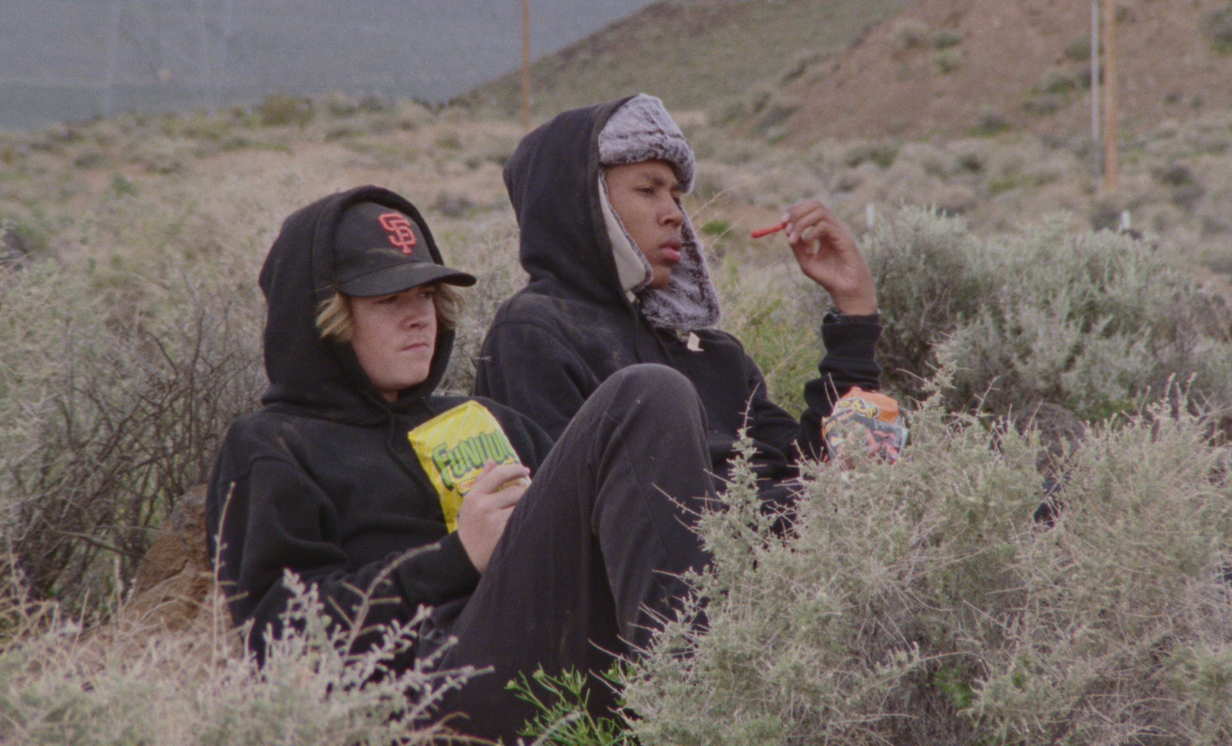Two people sitting in scrubland eating snacks. One wears a black cap, the other a hooded jacket and cap. Barren hills in background.