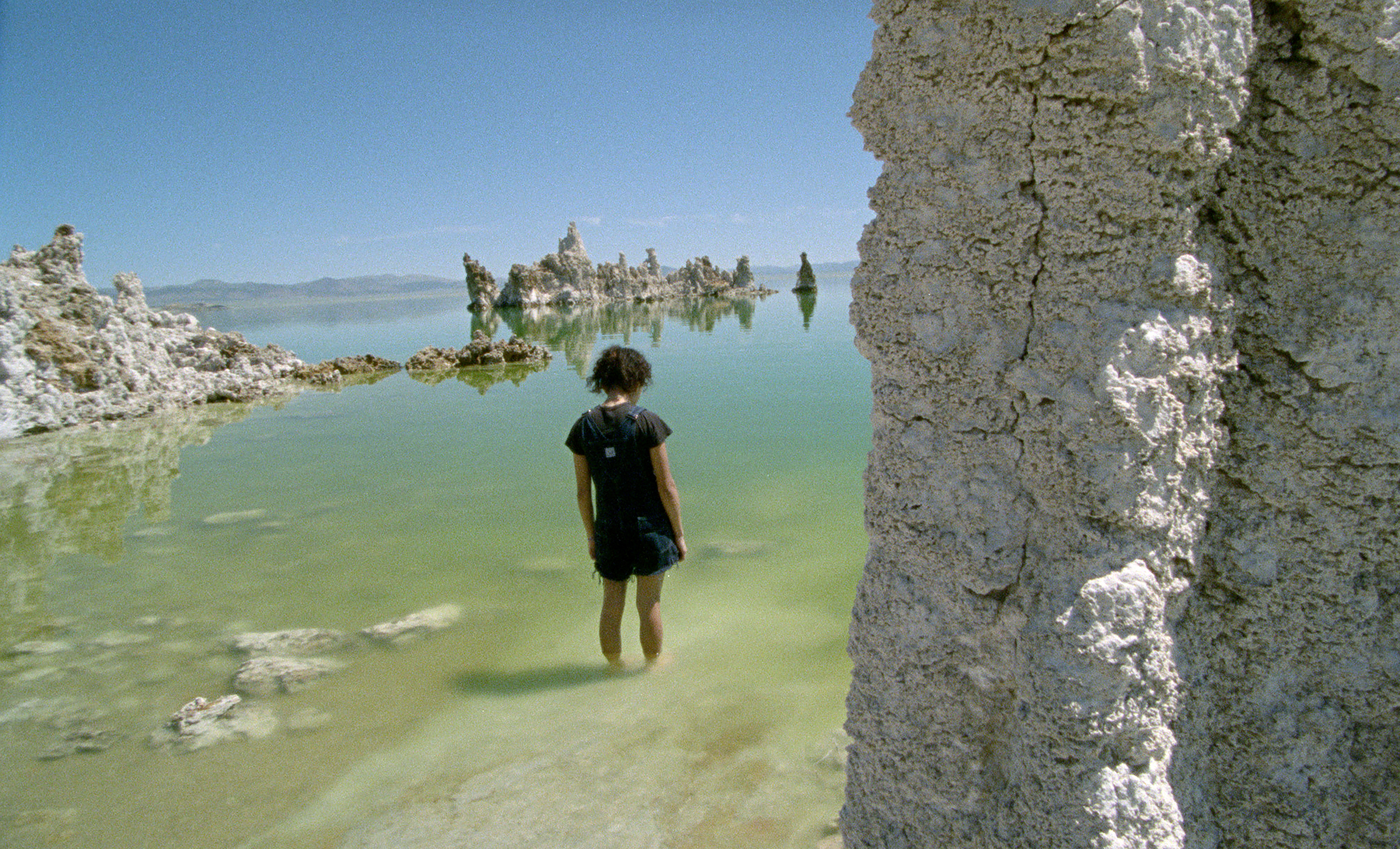 Person in dark clothing wading in shallow turquoise water amongst tall grey limestone formations under blue sky.