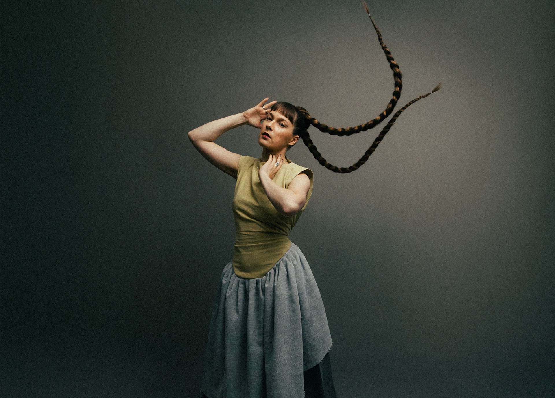 Woman with long dark braids in olive green top and blue skirt, hand shielding eyes, against grey-green gradient background.