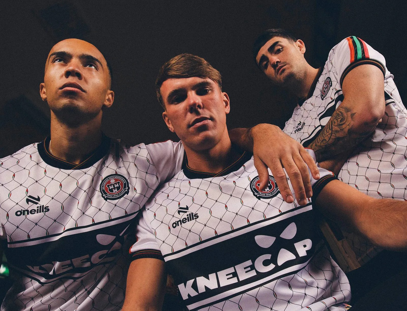 Three young men wearing white football jerseys with geometric pattern and "KNEECAP" text, posed together against dark background.