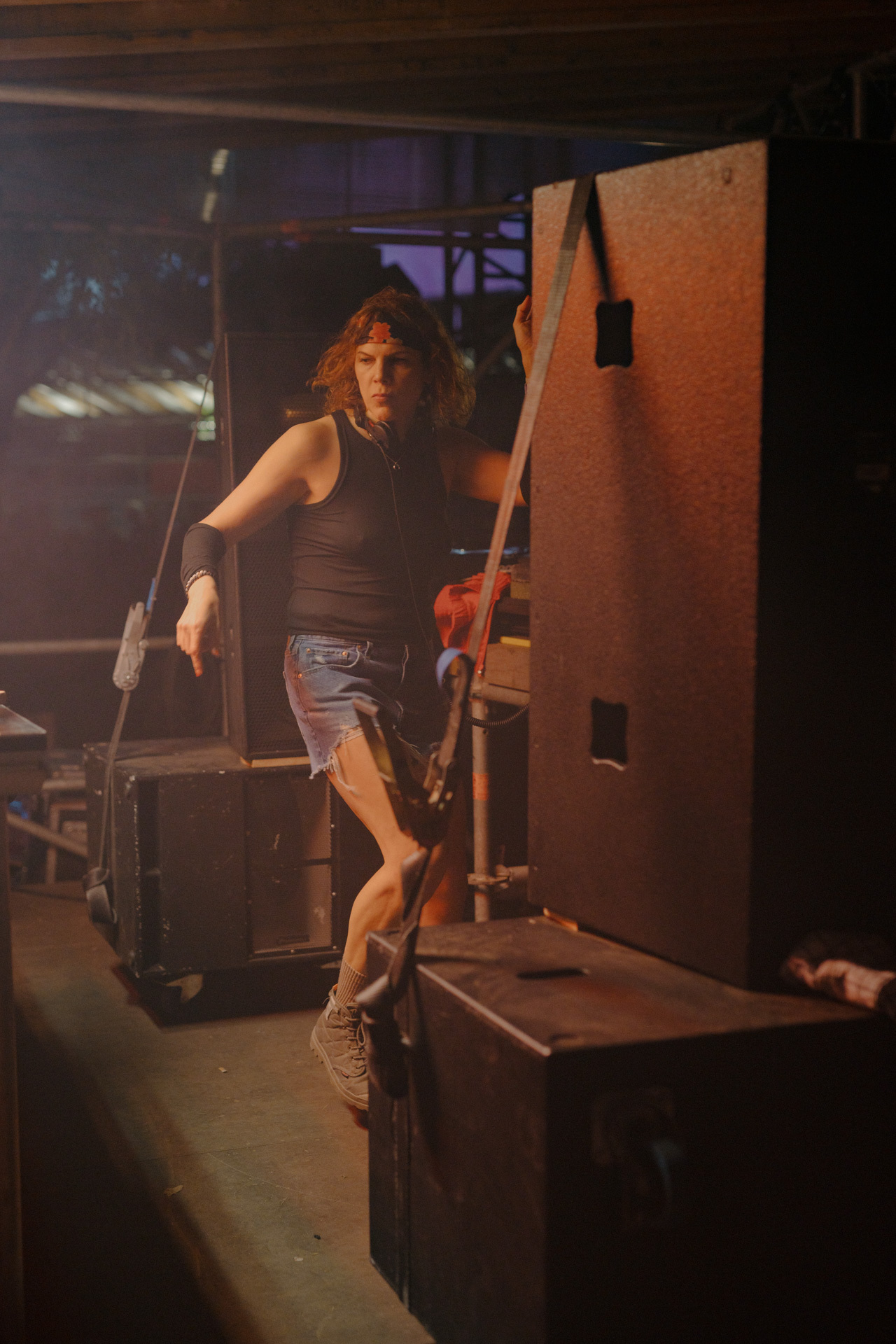 A person wearing dark clothing and holding a piece of equipment, standing in a dimly lit setting with various objects in the background.