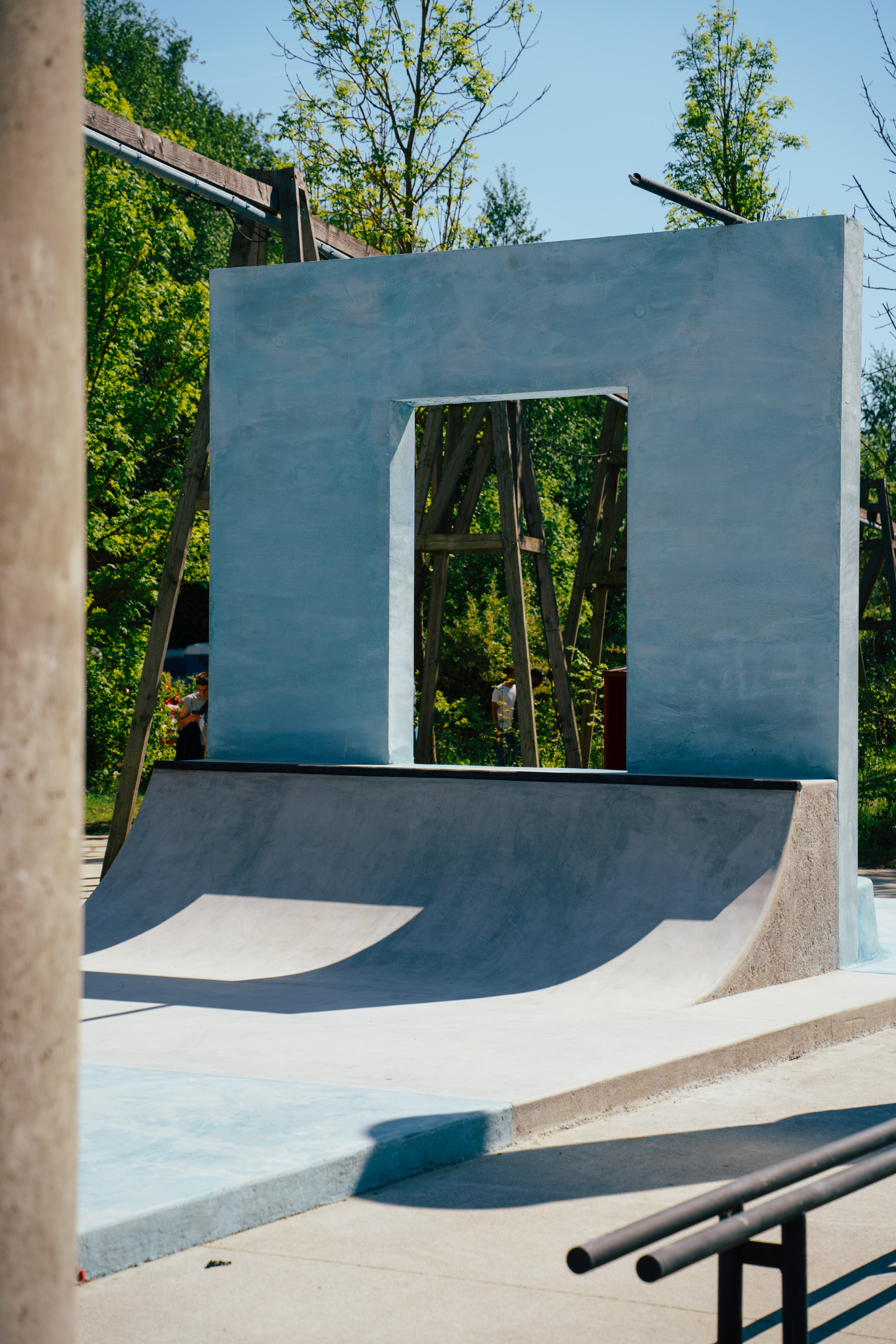 Concrete skate ramp with curved structures and reflective surfaces surrounded by trees.