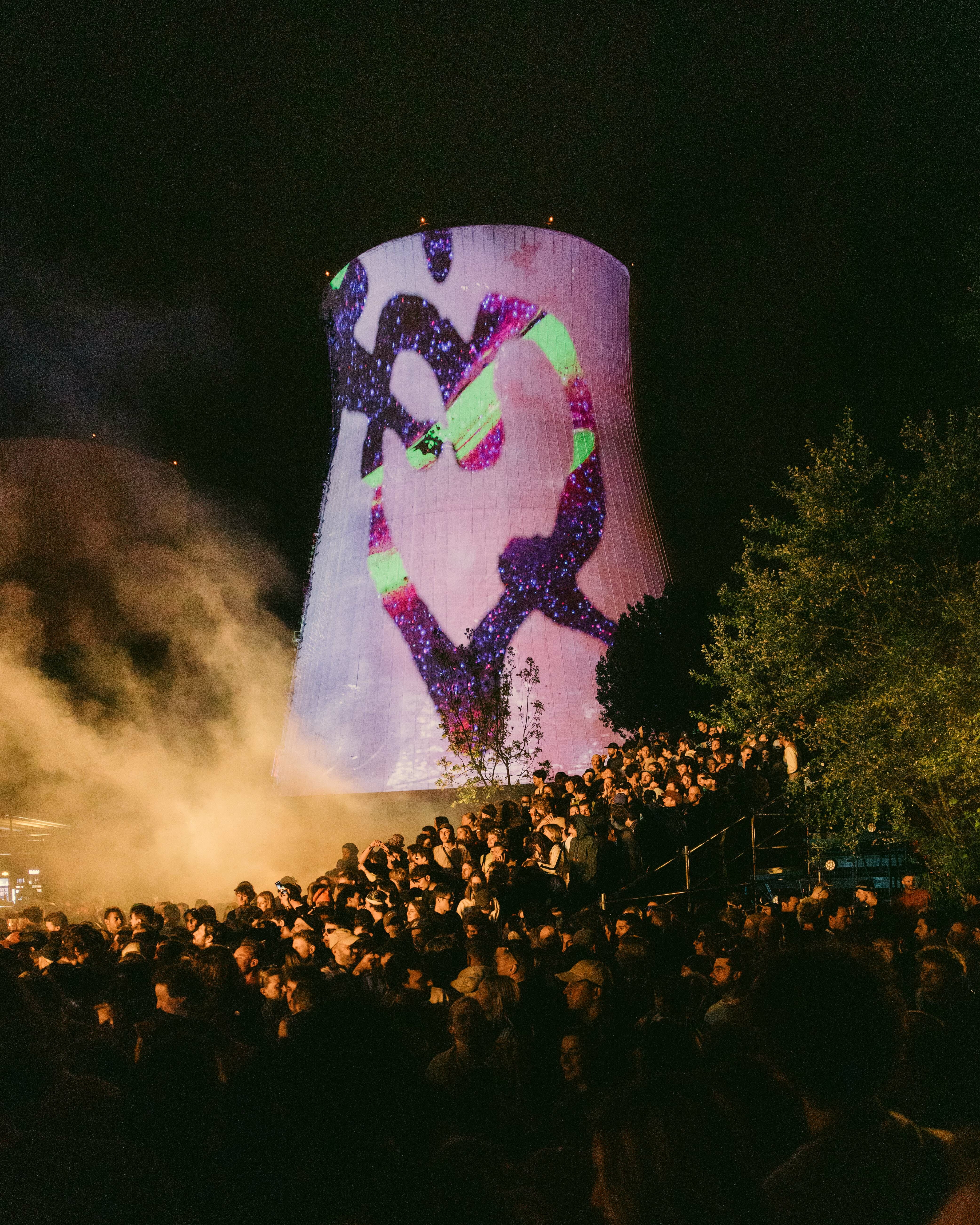 Brightly lit cylindrical structure with abstract shapes and figures projected onto it, surrounded by a large crowd of people.