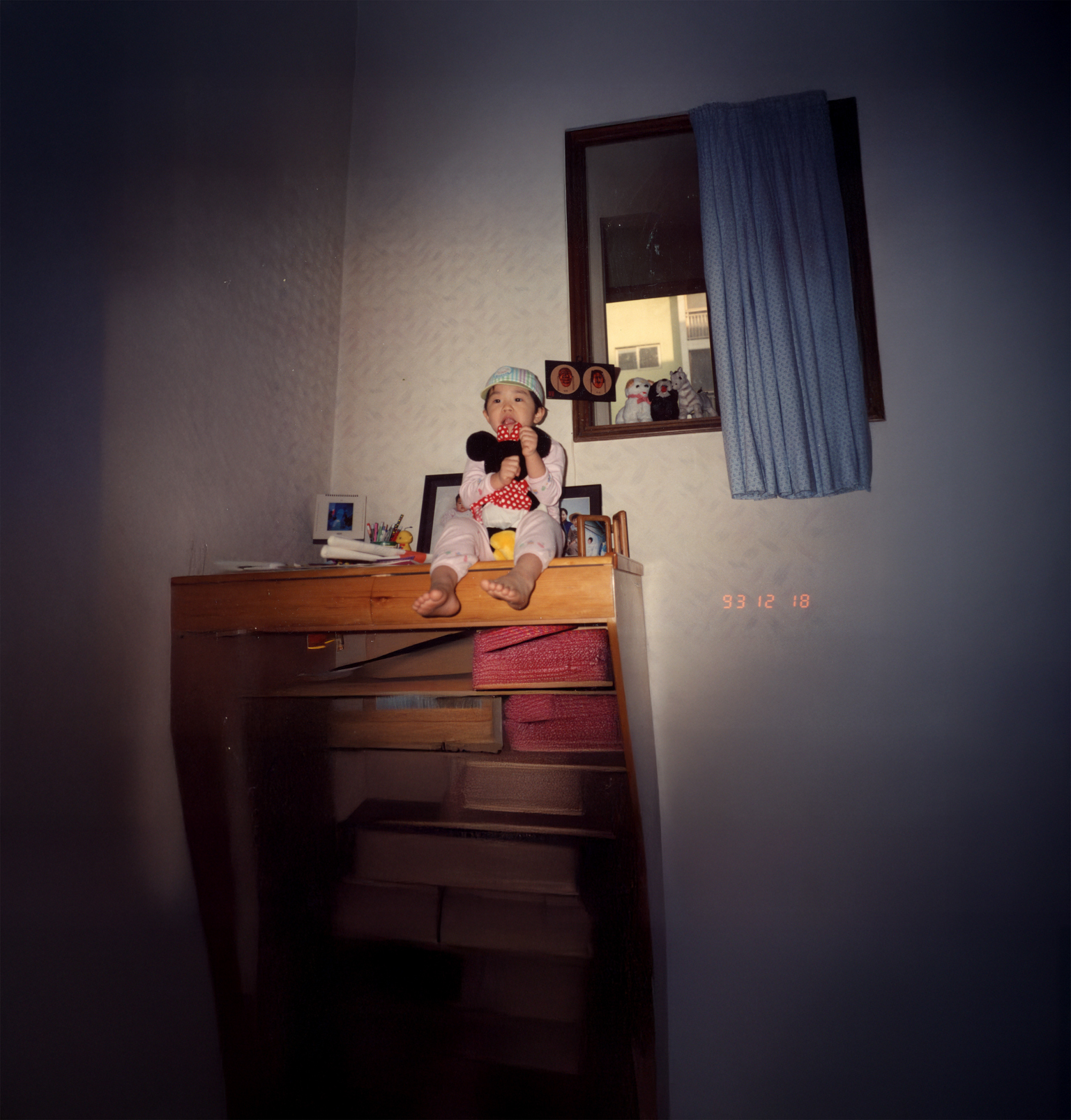 Child sitting on wooden chest of drawers in dimly lit bedroom, holding stuffed animal. Blue curtains visible in window above.