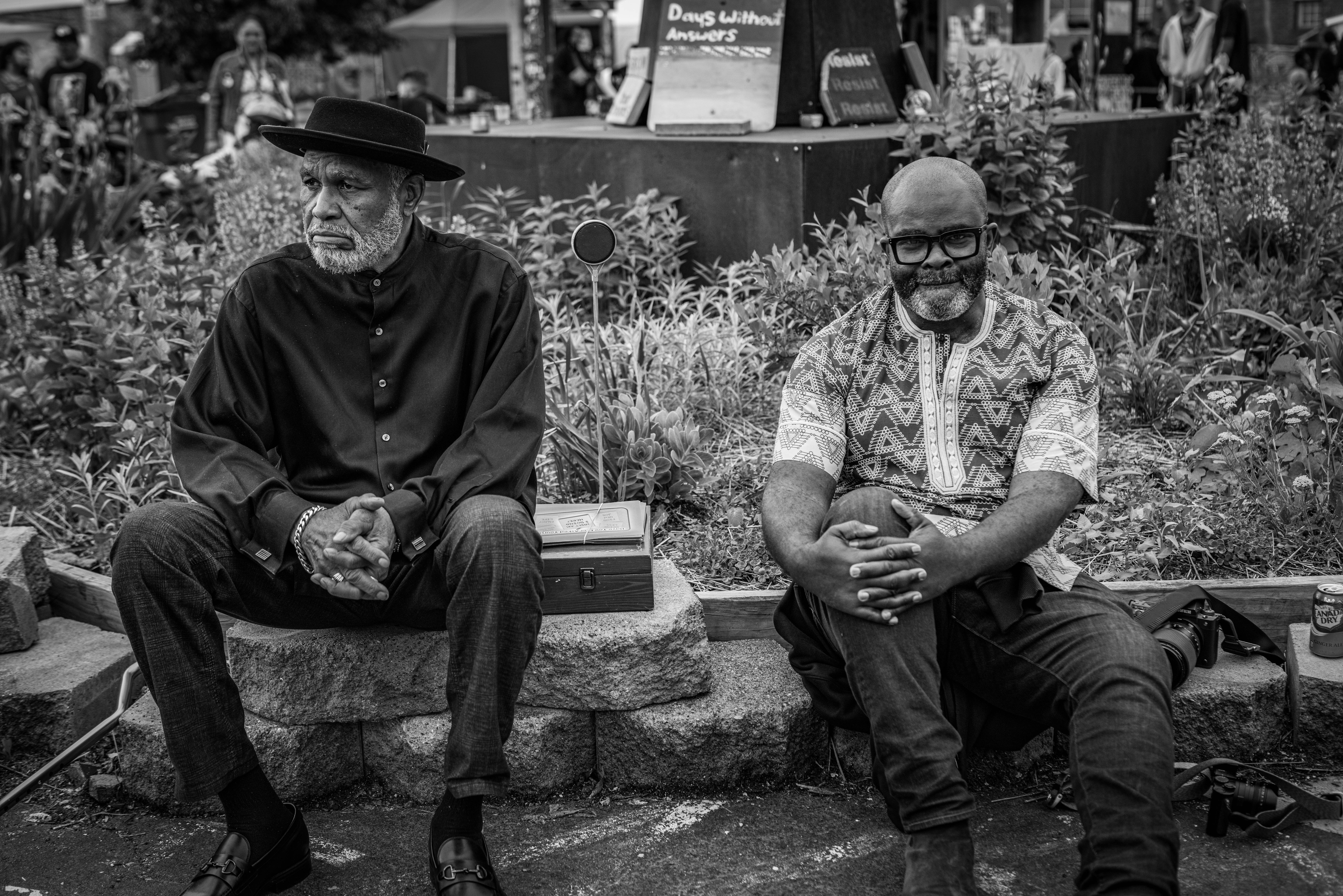 Two elderly men, one wearing a hat and the other with a beard, sitting on a bench in a garden setting with floral displays visible.