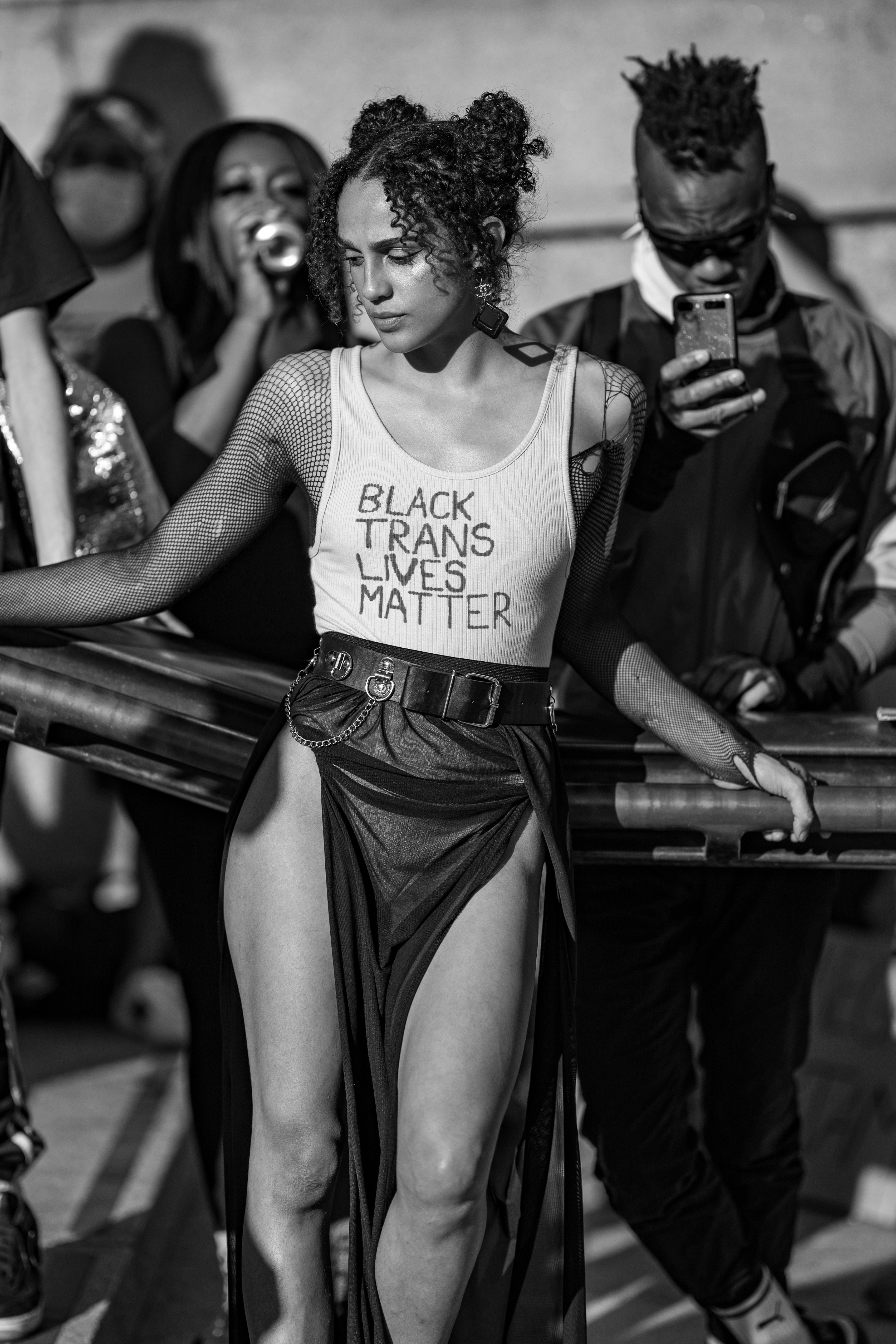 Black and white image of a person wearing a "Black trans lives matter" top standing among a crowd.
