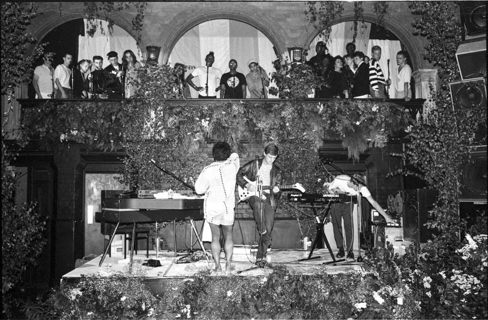 Black and white image showing musicians performing on outdoor stage with piano and equipment, audience watching from tiered stone balcony above.