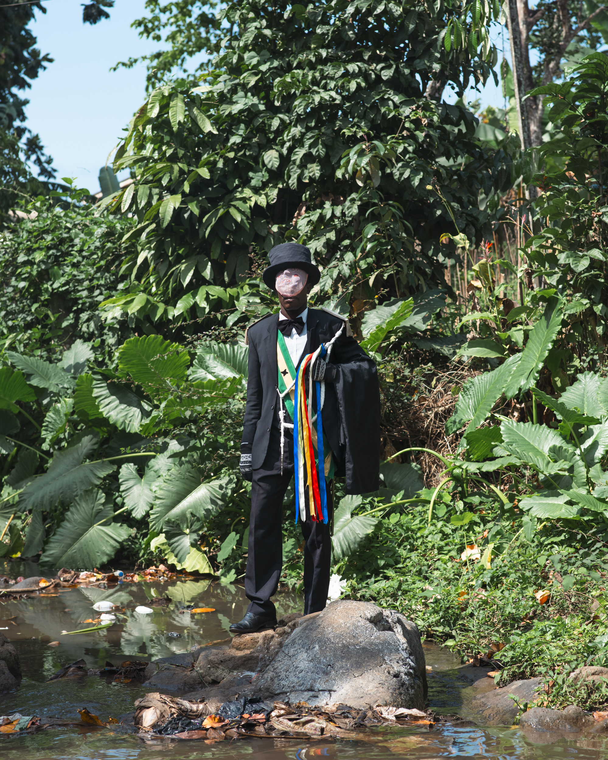Person in black graduation gown with colourful sash standing on rocks beside stream, surrounded by lush green tropical vegetation.