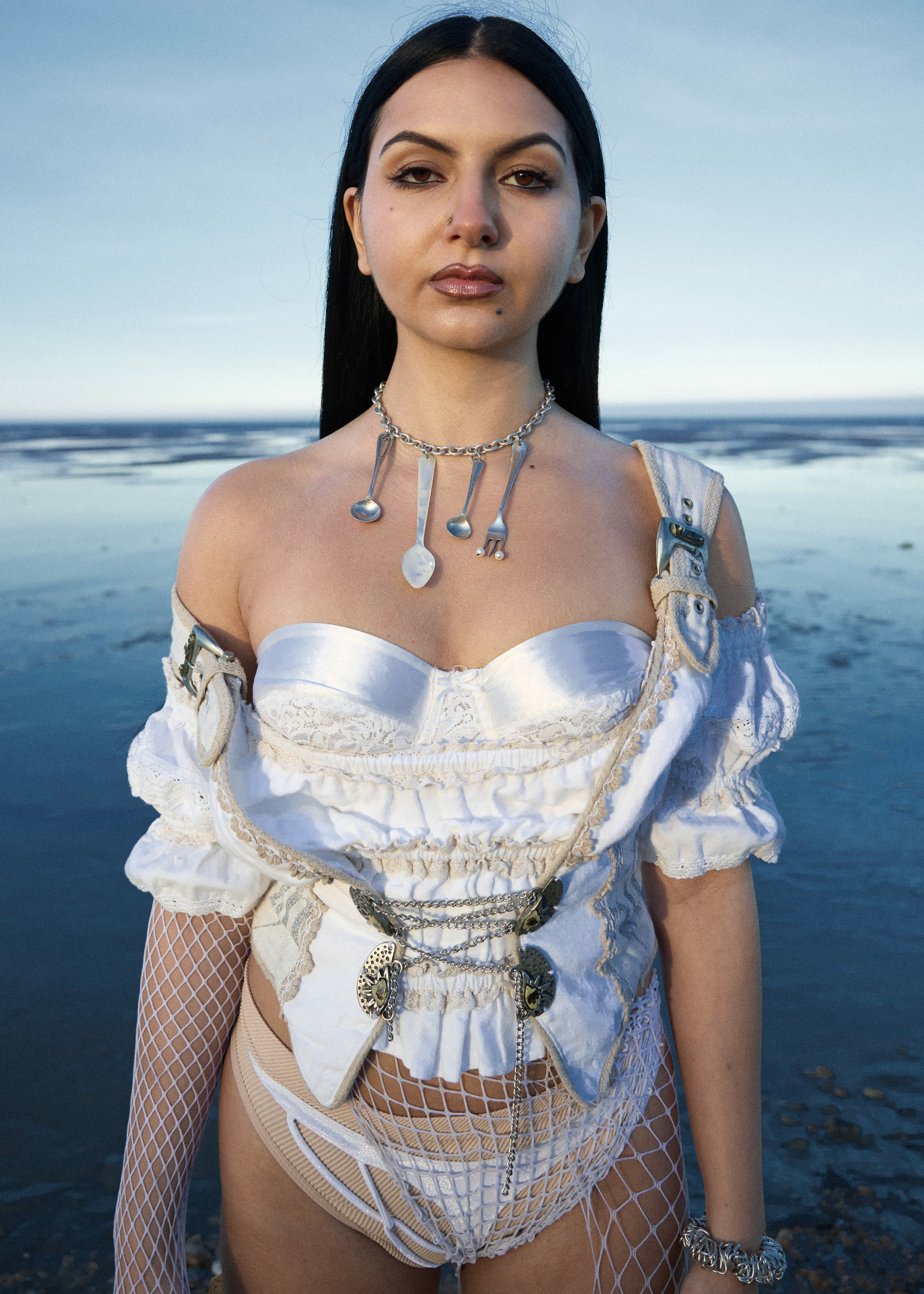 Woman wearing white lace dress and fishnet stockings, standing in front of ocean.