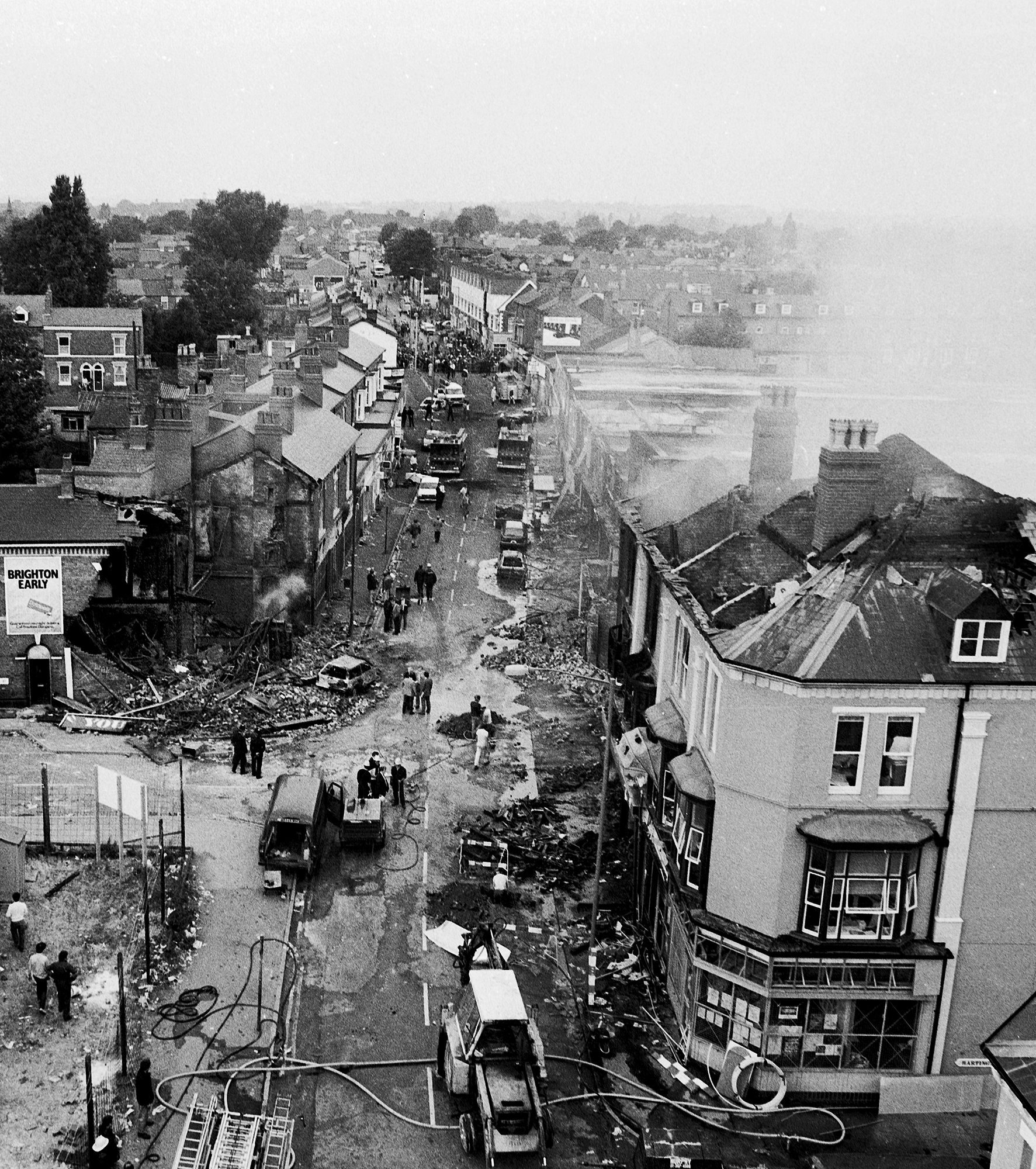 Black and white aerial view of bomb-damaged British town street with debris, emergency vehicles, and people amongst destroyed buildings.