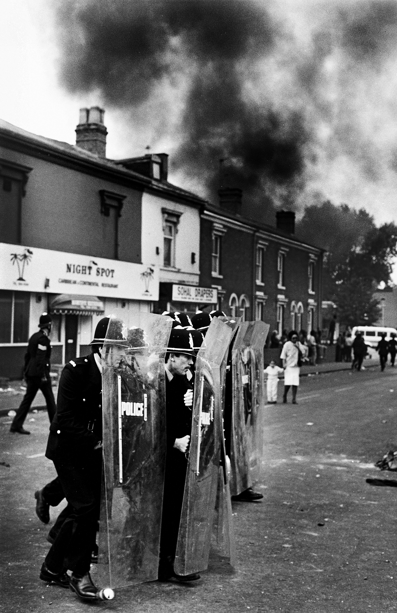 Police officers with riot shields on street during civil unrest, black smoke billowing above terraced houses and shops.
