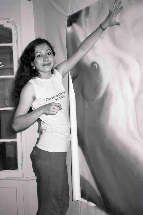 Woman in white t-shirt and dark trousers holds onto vertical pole with one hand, other arm raised, shadow cast on wall behind.