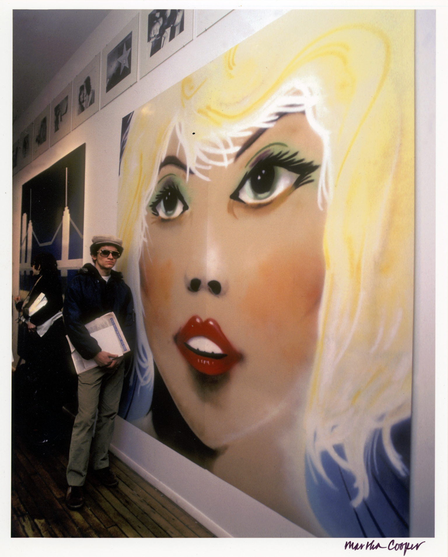 Large wall mural of blonde woman's face with red lips and green eyes; person in black coat and sunglasses standing beside it.