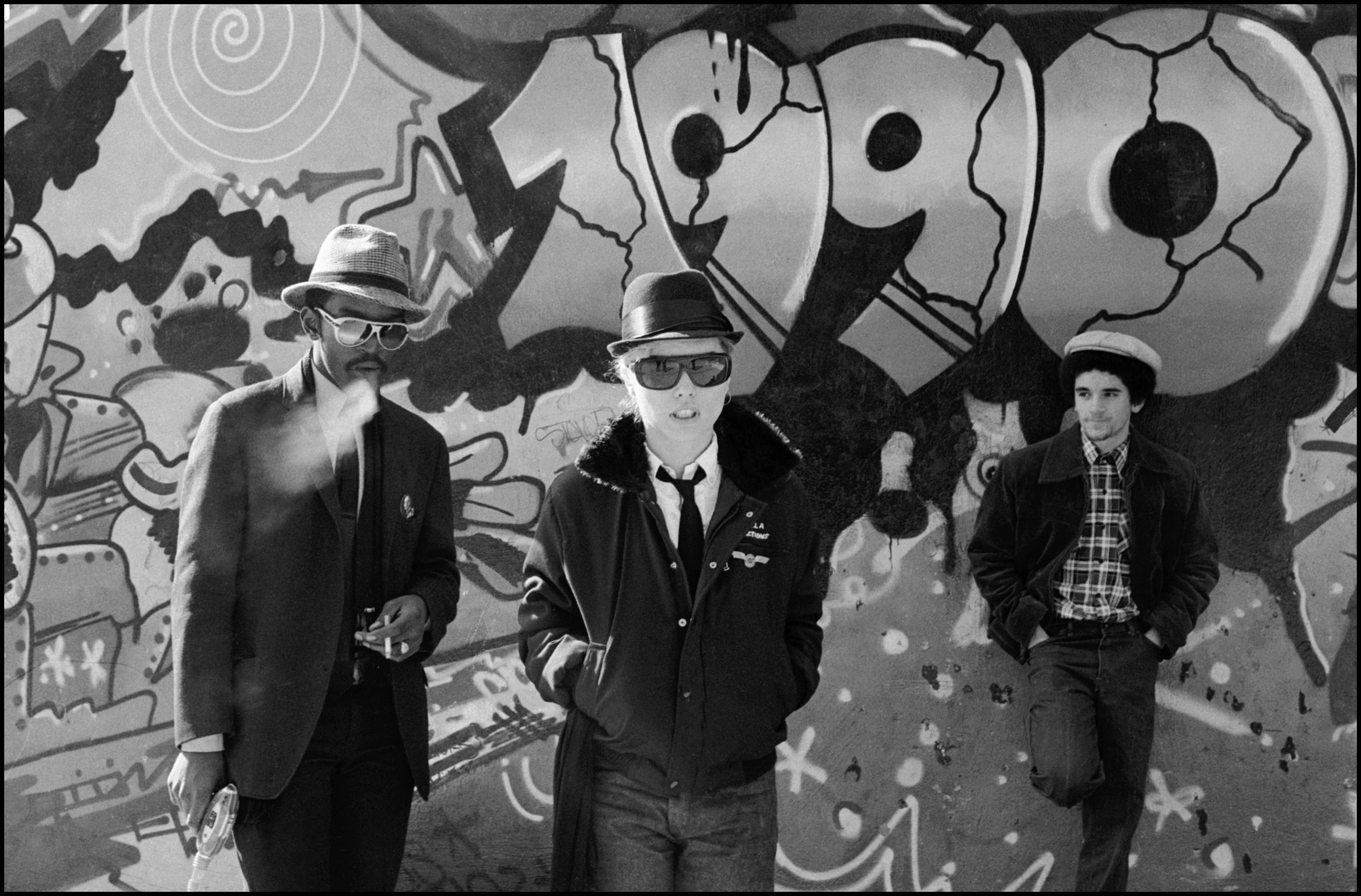 Three men in hats and sunglasses standing against colourful graffiti wall with cartoon-style characters and swirling patterns.