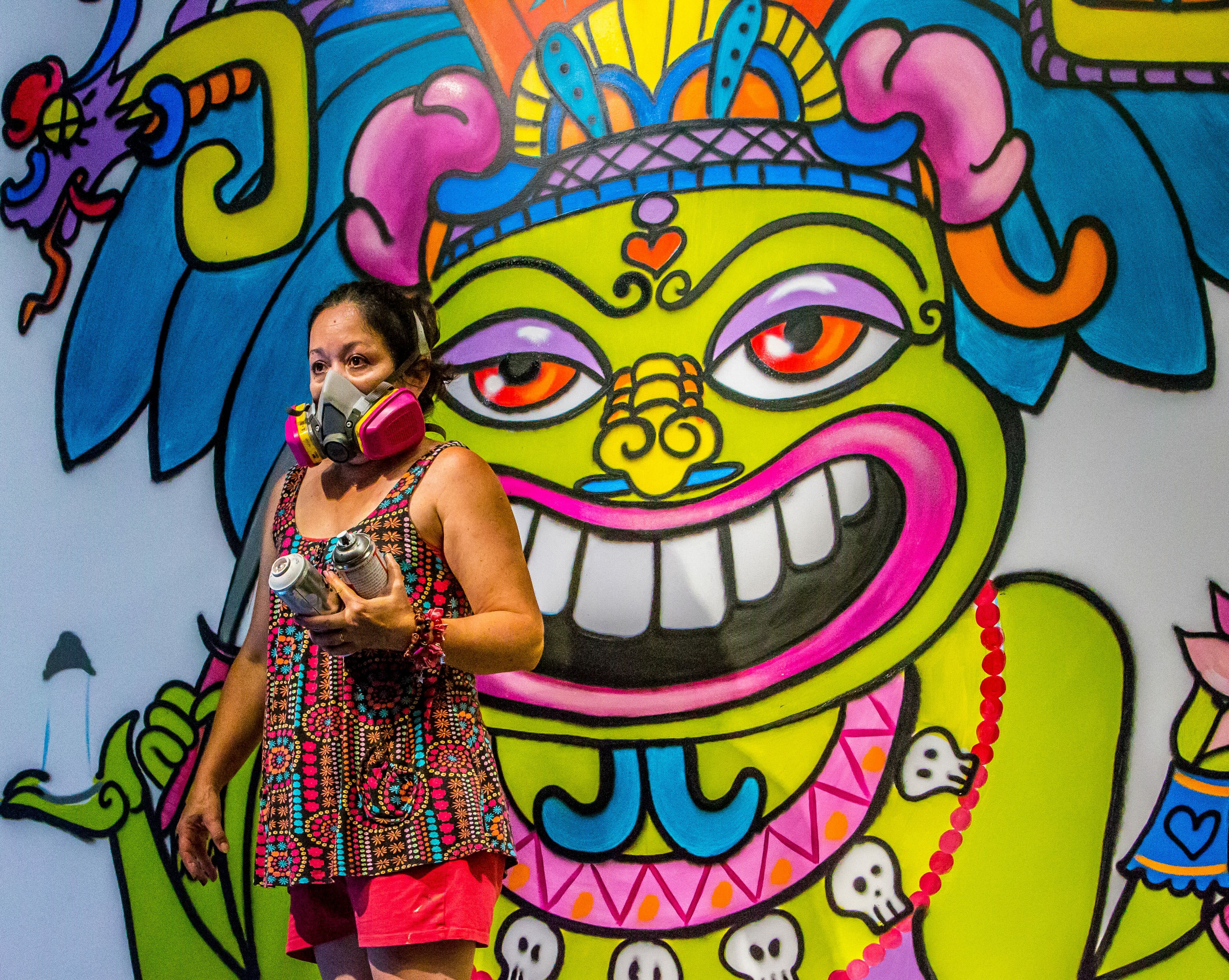 Woman in patterned dress and face mask standing before vibrant street art mural featuring green mythical creature with orange eyes, white teeth, and colourful geometric patterns.