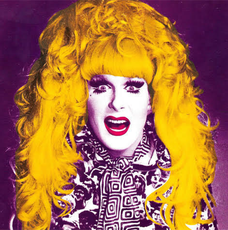 Close-up of a person with long, curly blonde hair, wearing bold make-up and an ornate, patterned outfit.