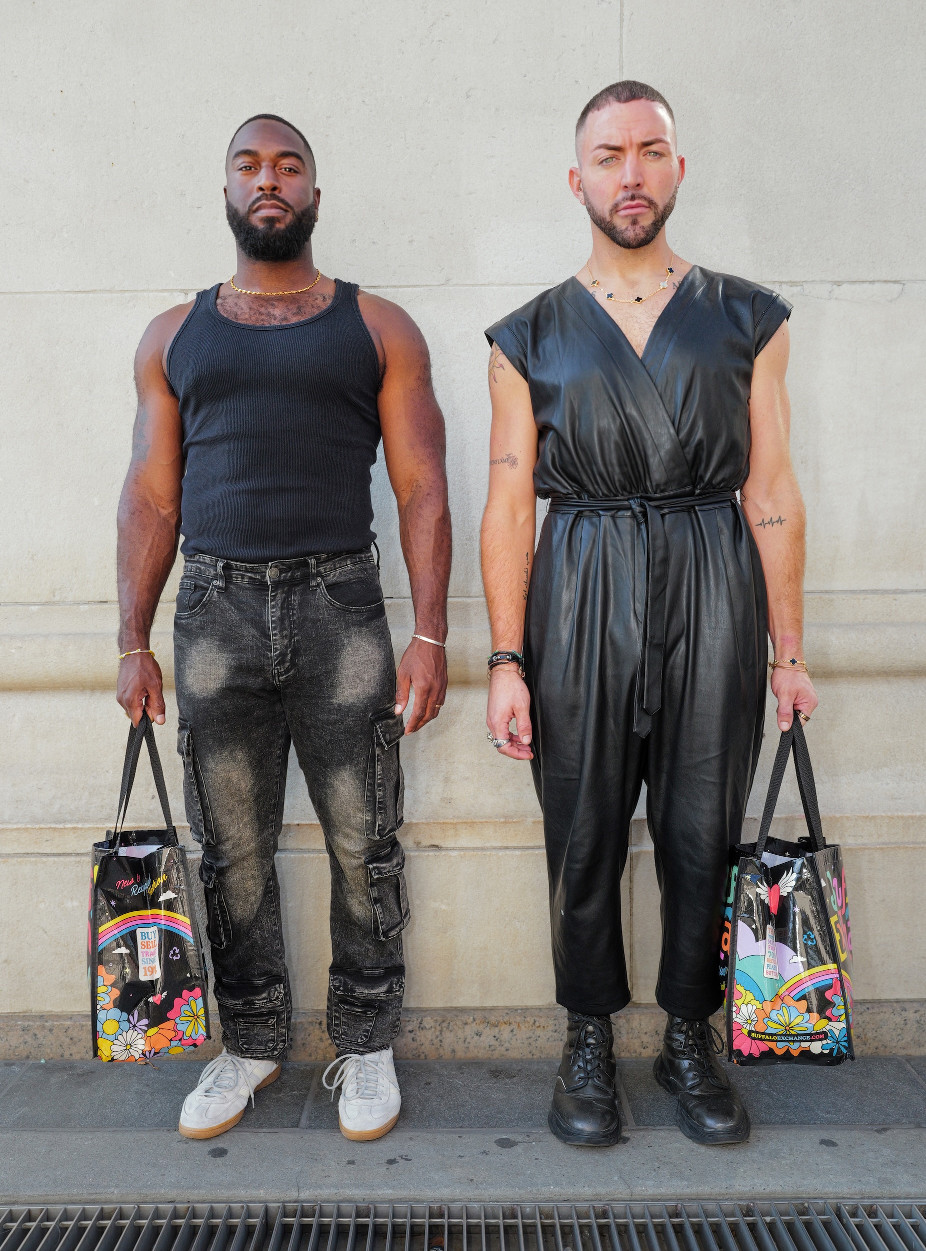 Two men standing against white wall. Left: Black man in dark vest and distressed jeans. Right: White man in black leather jumpsuit. Both holding colourful patterned bags.