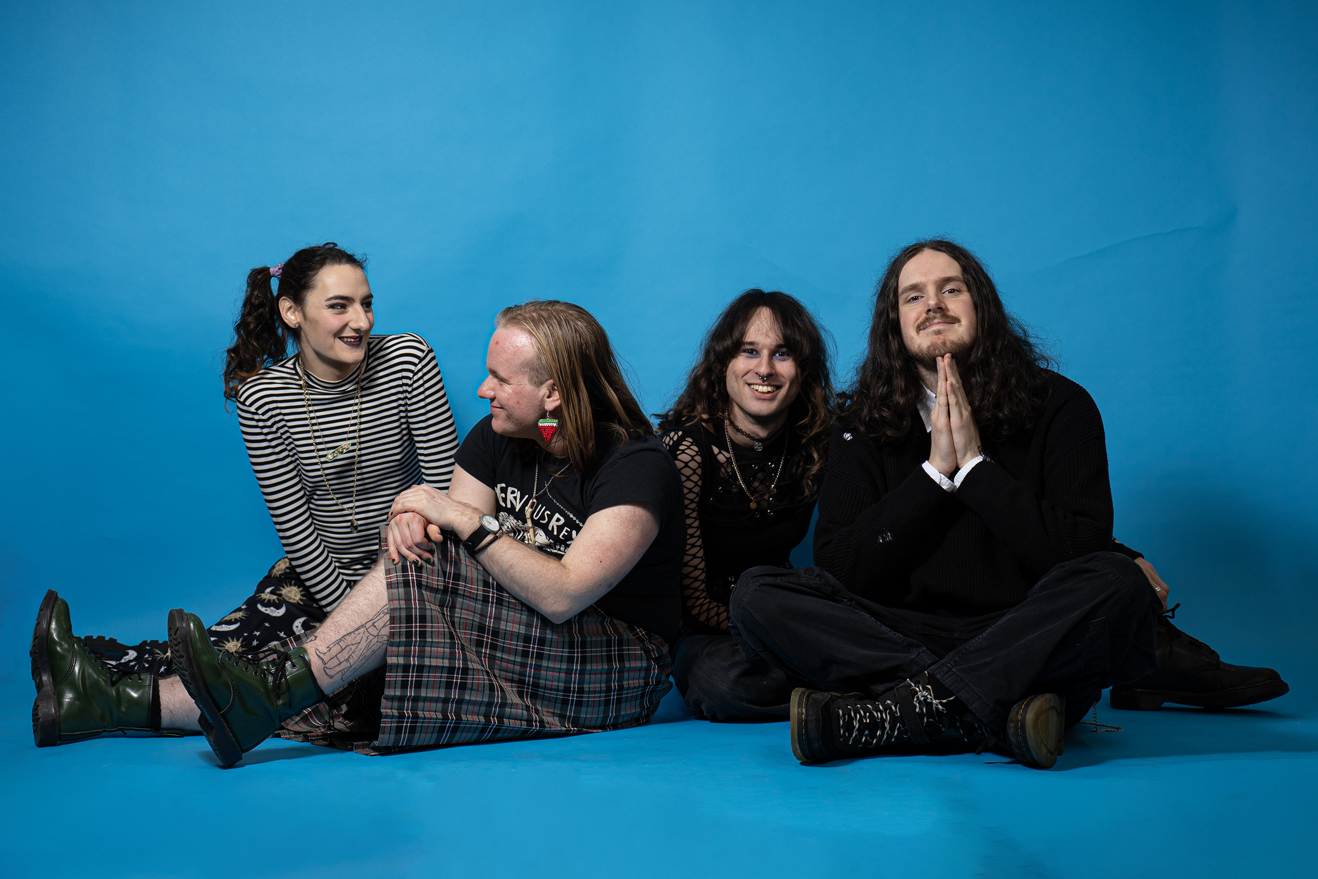 Four people sitting together against bright blue backdrop, wearing casual dark clothing including stripes and plaid patterns.