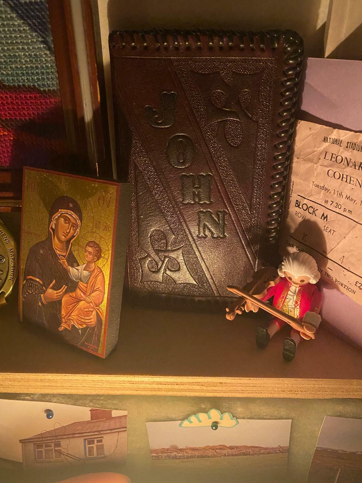 Ornate leather-bound book with embossed religious symbols, framed religious icon, decorative trinkets, and art prints on a wooden shelf.