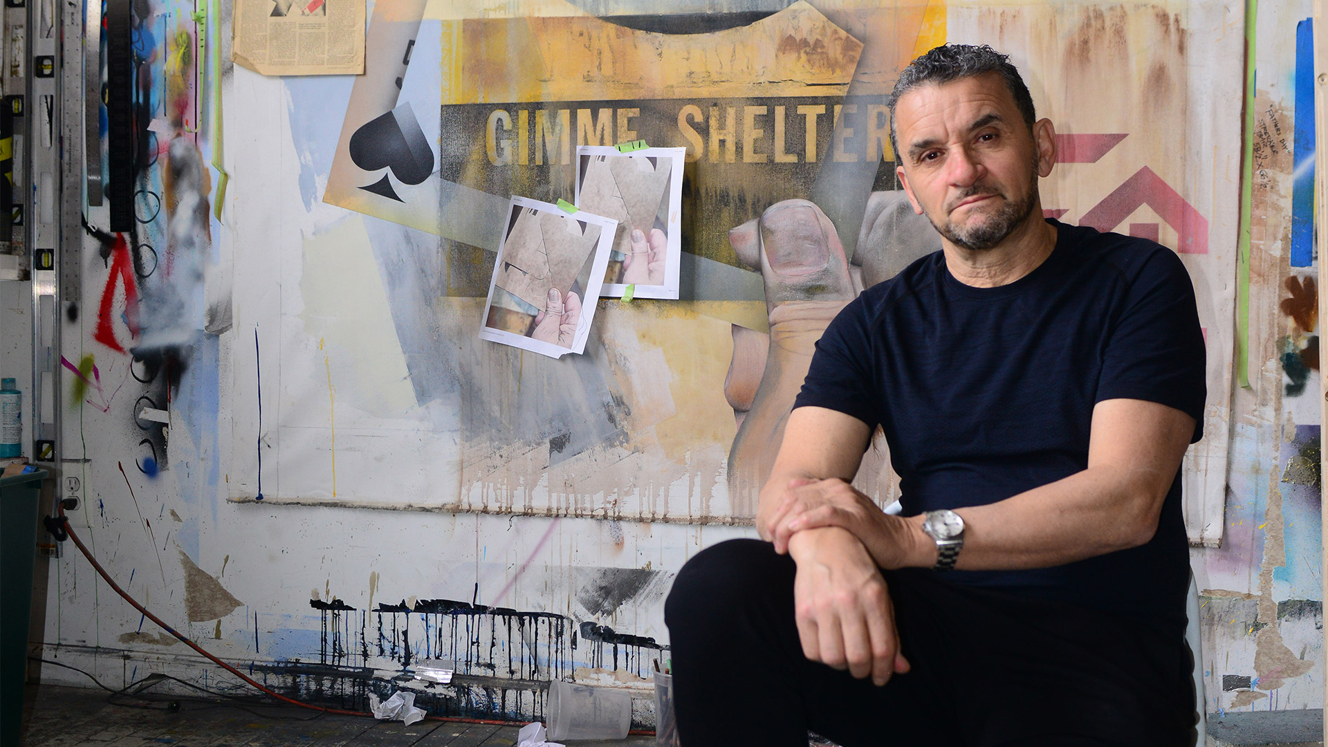 Man in black t-shirt sitting in art studio with colourful abstract paintings and mixed media works on walls behind him.