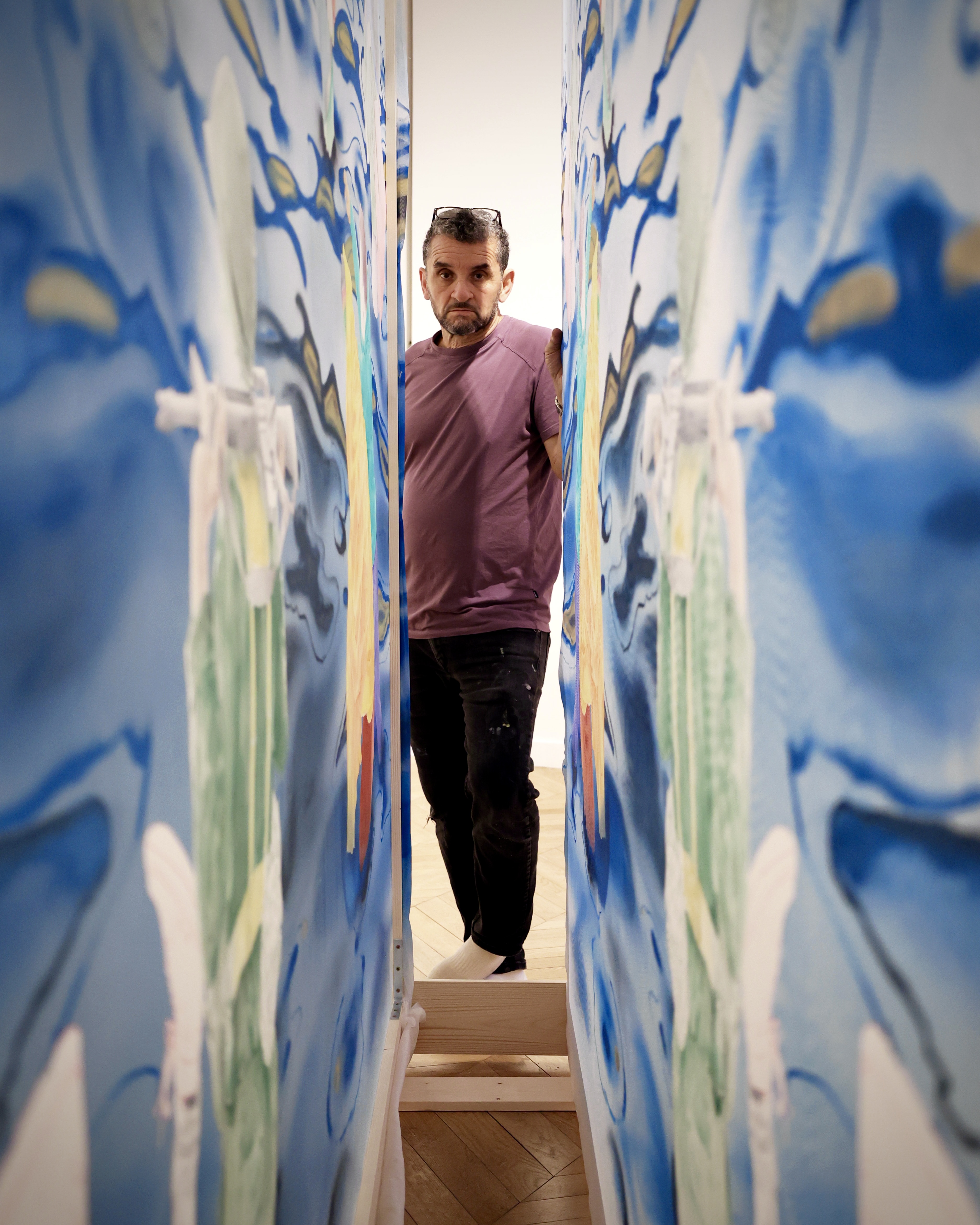 Man in burgundy jumper standing in narrow corridor with blue, white and green painted walls creating tunnel-like perspective.