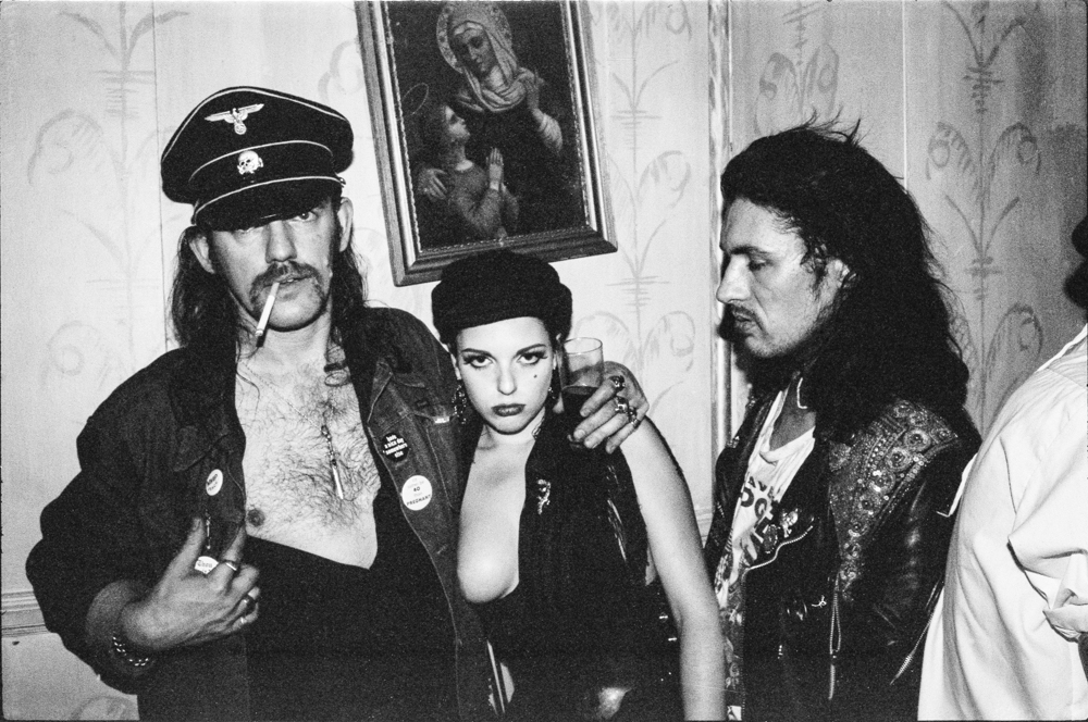 Three people in black leather outfits posing indoors. Left person wears military-style cap, centre person in low-cut top, right person has long dark hair.