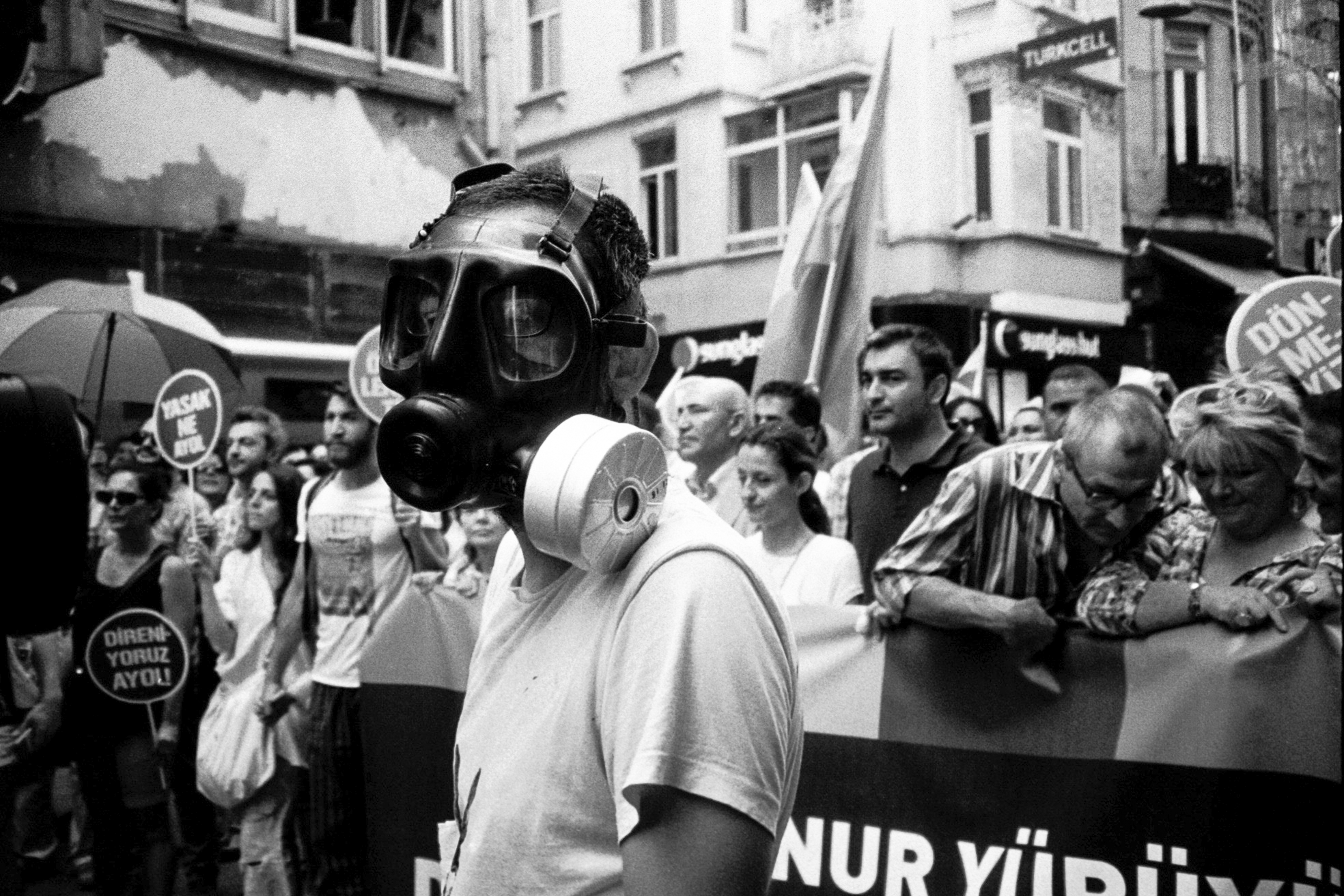 Crowd of people wearing gas masks and protest banners in black and white image.