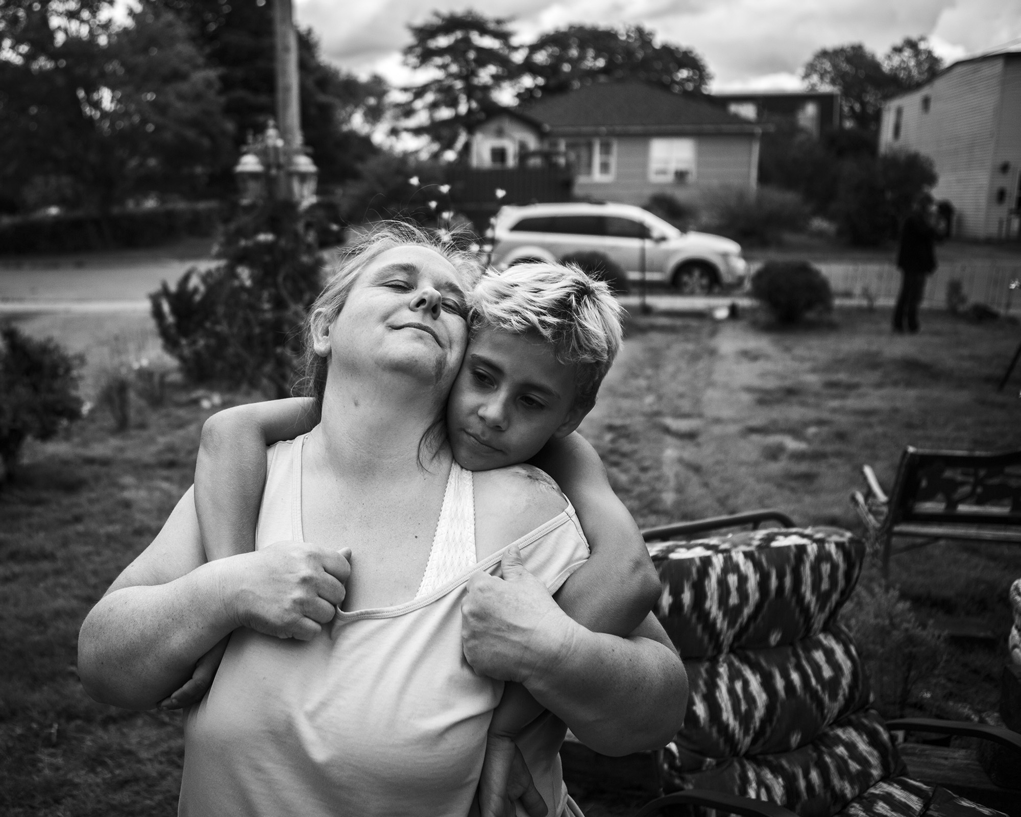Two people, a woman and a young child, embrace in a black and white outdoor scene with a house and car in the background.