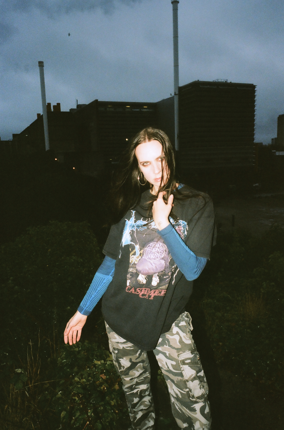 Person with long dark hair wearing black graphic t-shirt and blue sleeves, camouflage trousers, standing outdoors at dusk with industrial buildings behind.
