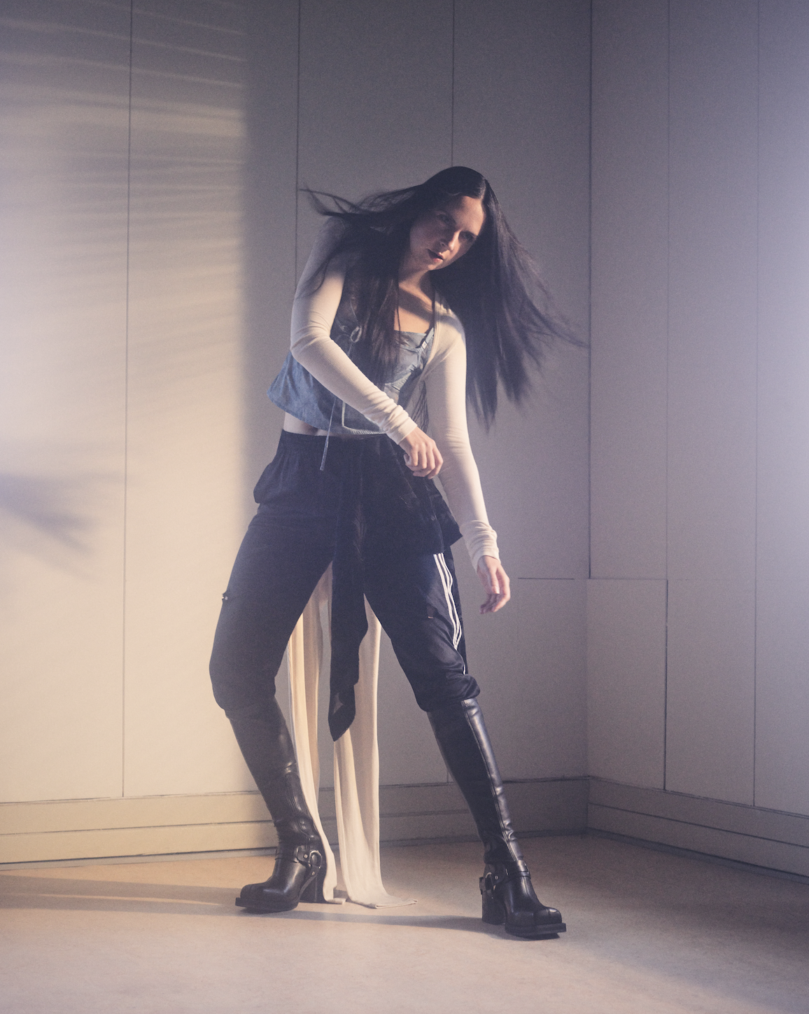 Woman with long dark hair in grey top, black trousers and boots, standing in corner with grey walls and warm lighting.