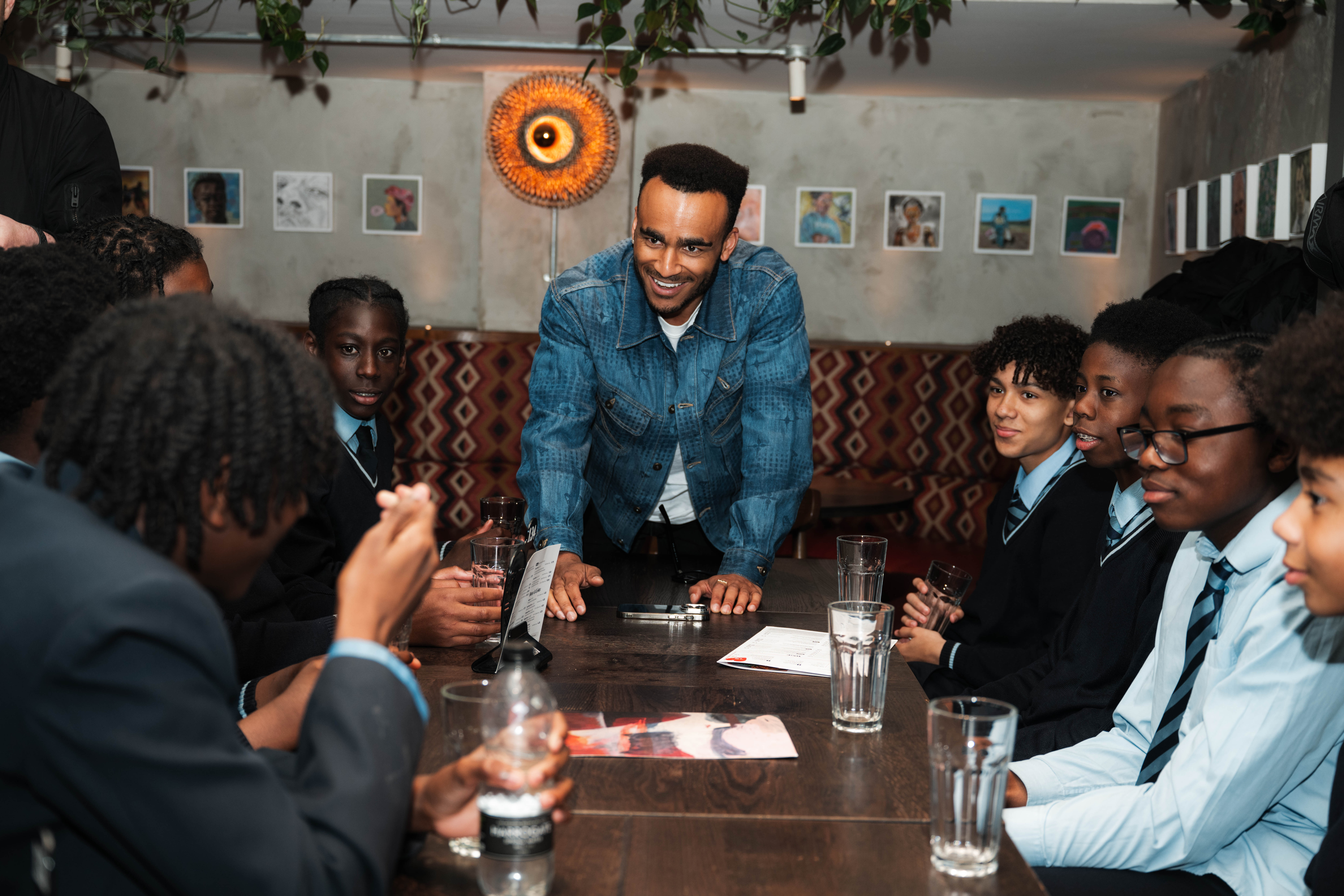 Man in denim jacket standing at conference table surrounded by seated colleagues in business attire in restaurant setting.